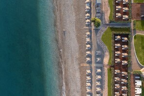 An aerial view of a beachside resort with rows of umbrellas and lounge chairs, a wooden walkway, and a paved path along the beach.