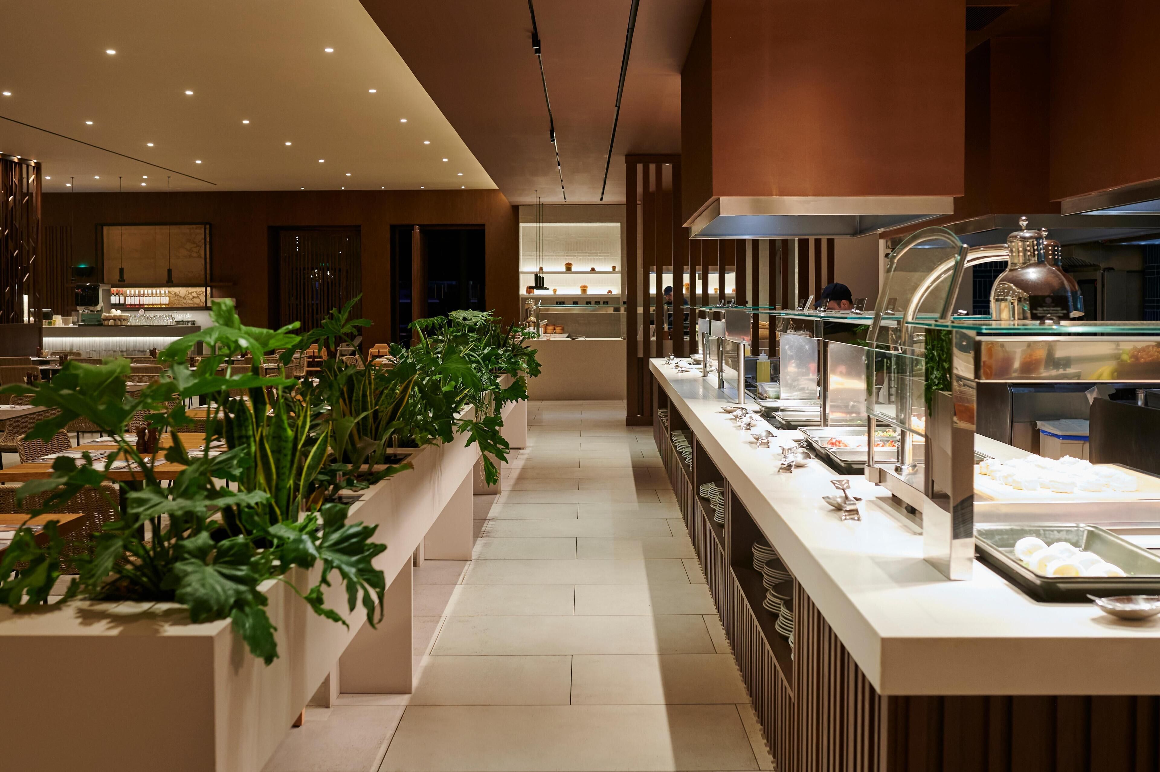 Un moderno vestíbulo de hotel con un mostrador de recepción alargado, una zona de buffet con diversas opciones de comida y un área de estar con plantas.
