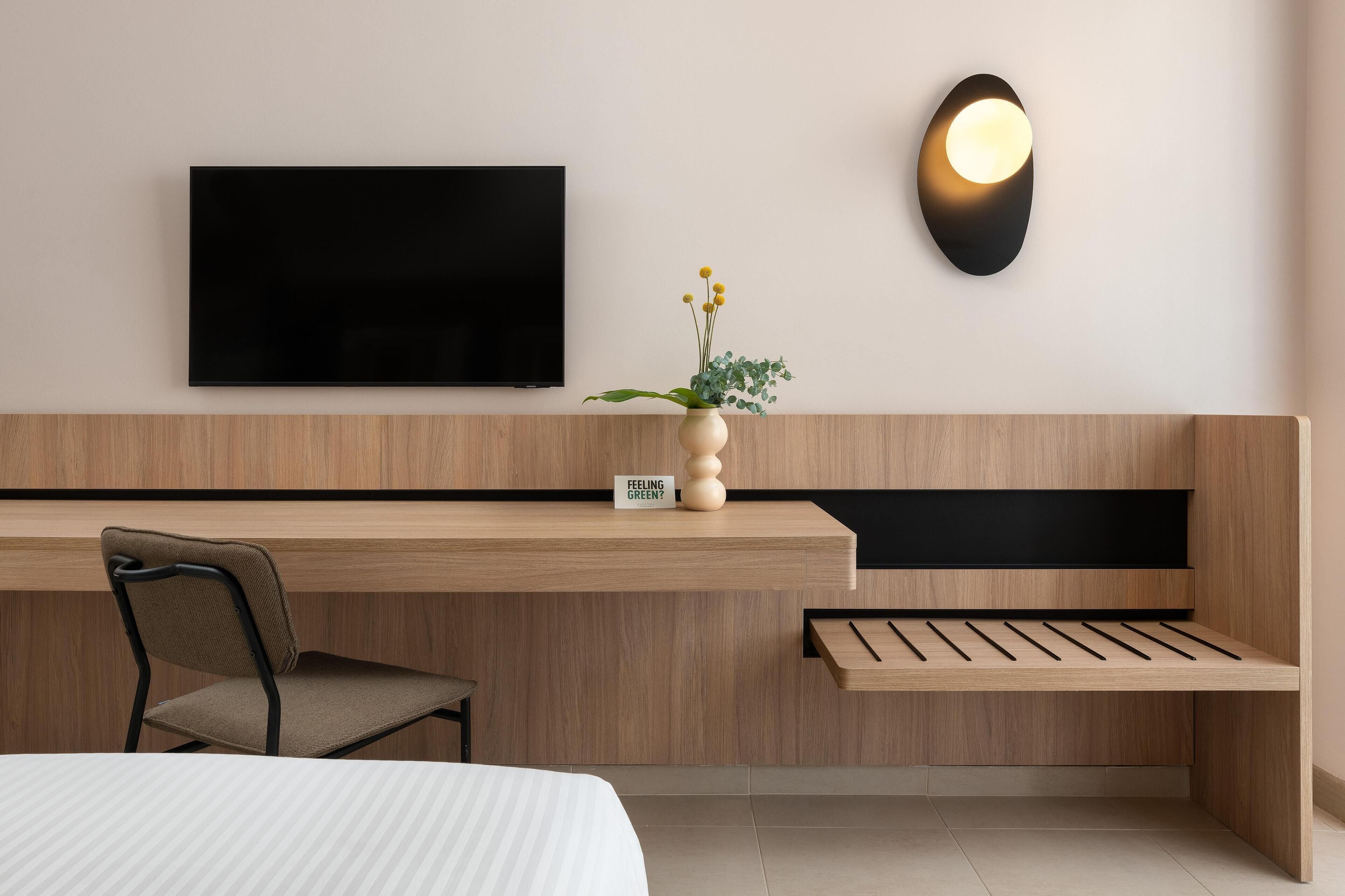 Ett modernt hotellrum med en receptionsdisk i trä, en platt-TV som är monterad på väggen, en liten krukväxt och en stol.