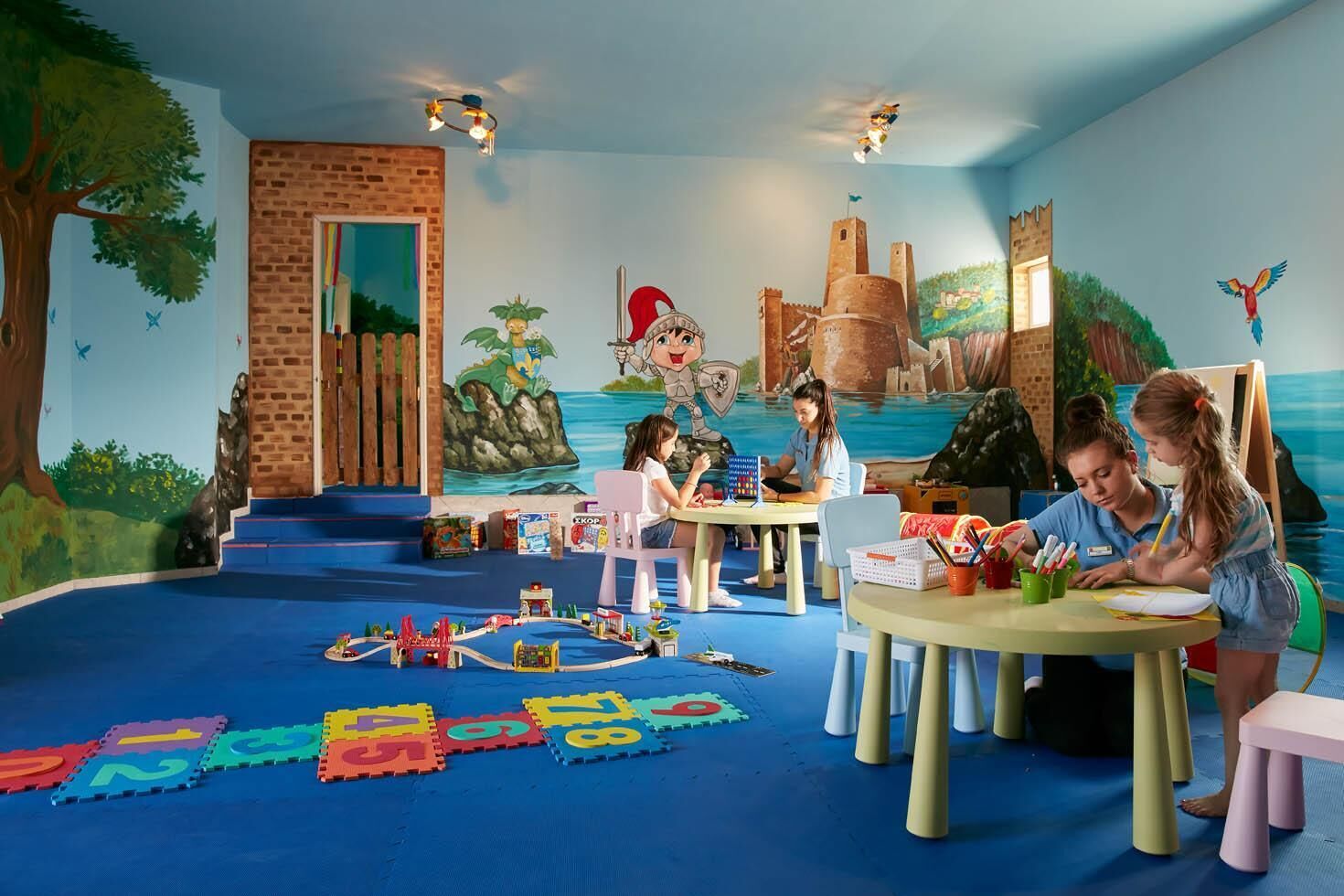 Uma sala de jogos infantil com brinquedos educativos, um mural de castelo e um tapete azul.