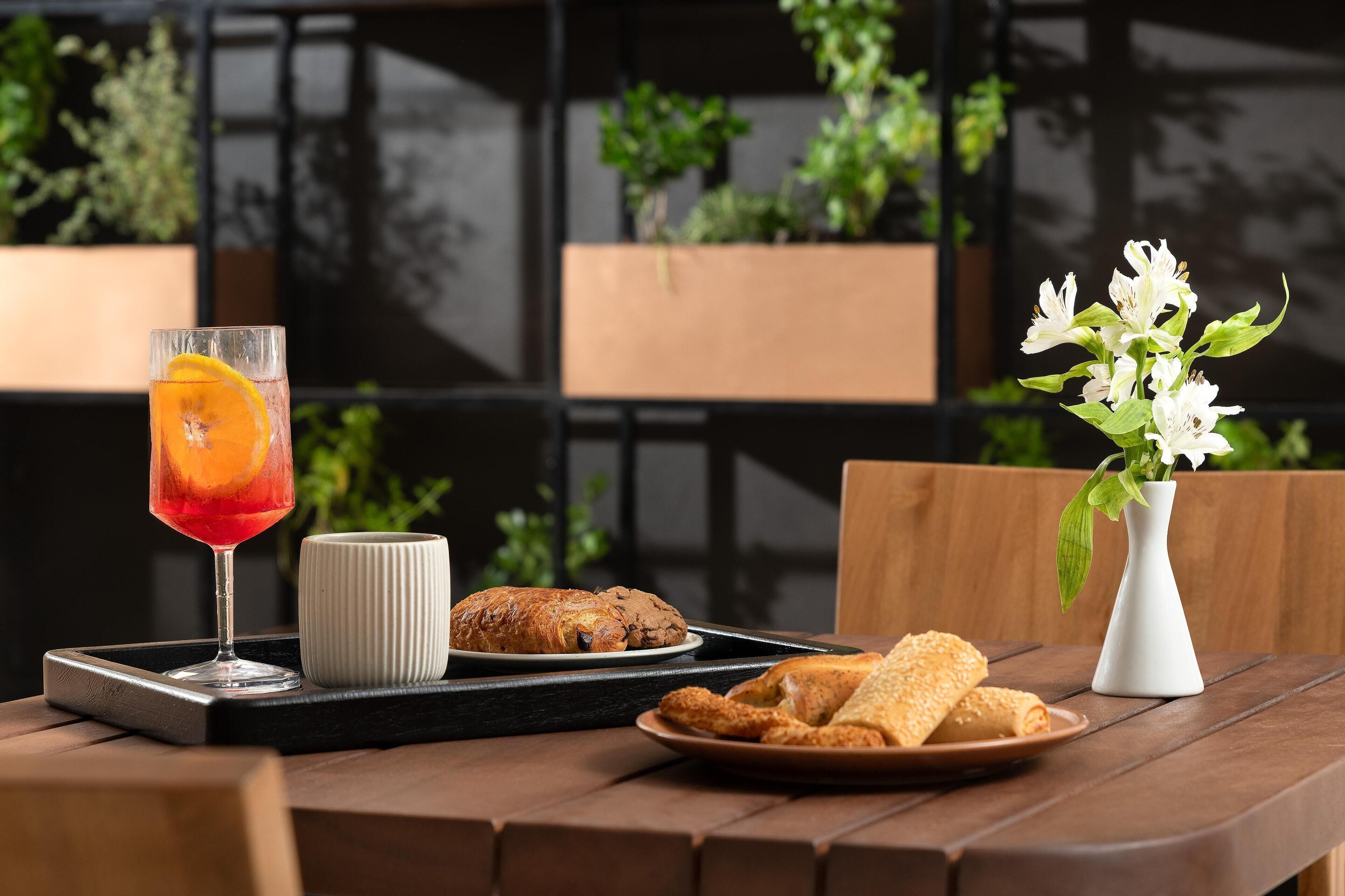 A table with a glass of red drink, a white mug, a plate of croissants, and a vase with white flowers.