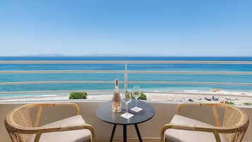 Un balcón con dos sillas de mimbre y una mesa, con vista a una playa de aguas azules cristalinas y montañas lejanas.