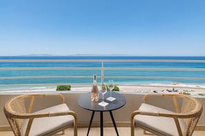 A balcony with two wicker chairs and a table, overlooking a beach with clear blue waters and distant mountains.