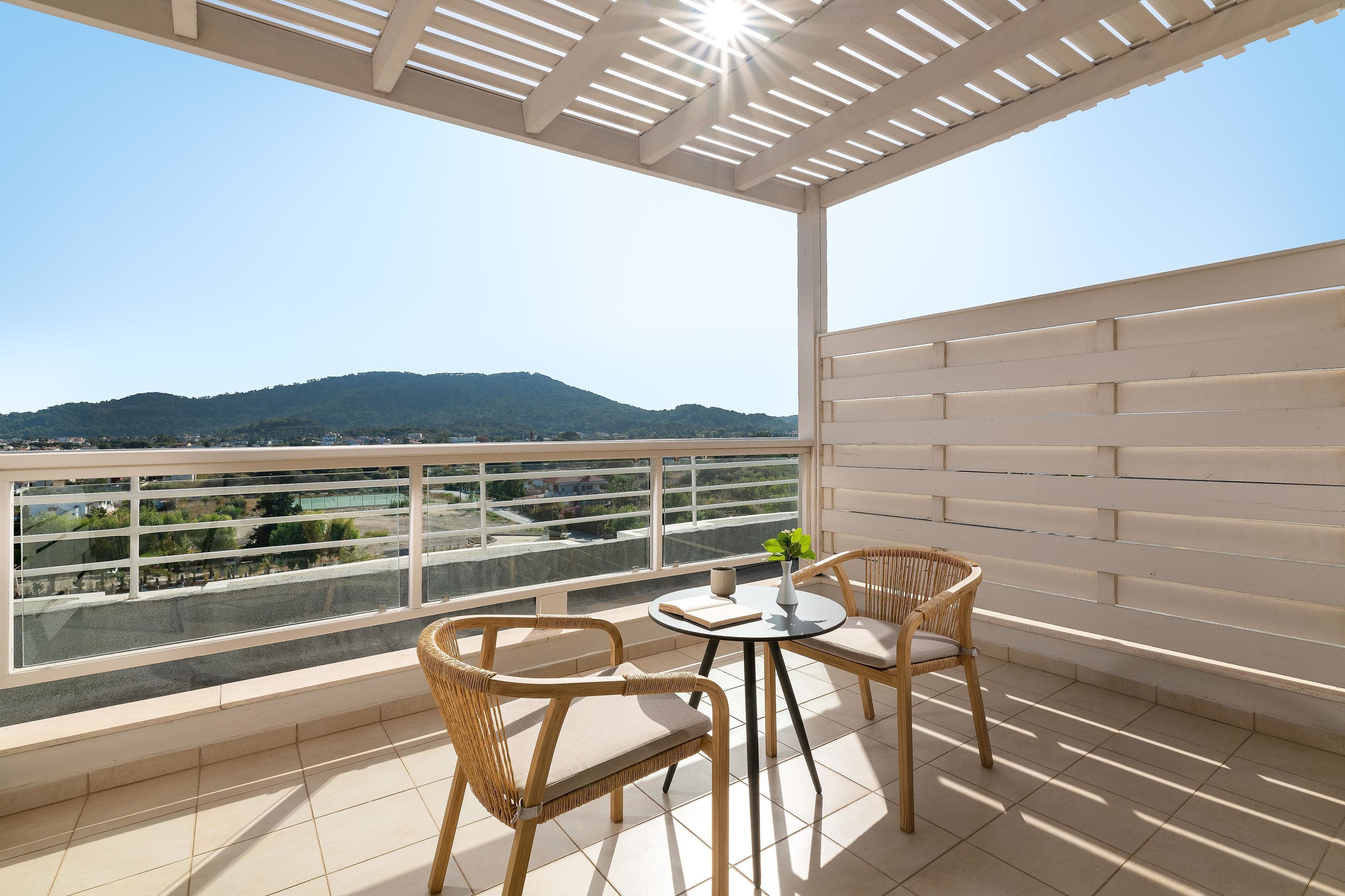 Un balcón con vistas a la montaña, dos sillas de mimbre, una mesita con un florero y un libro, y una estructura de pérgola.