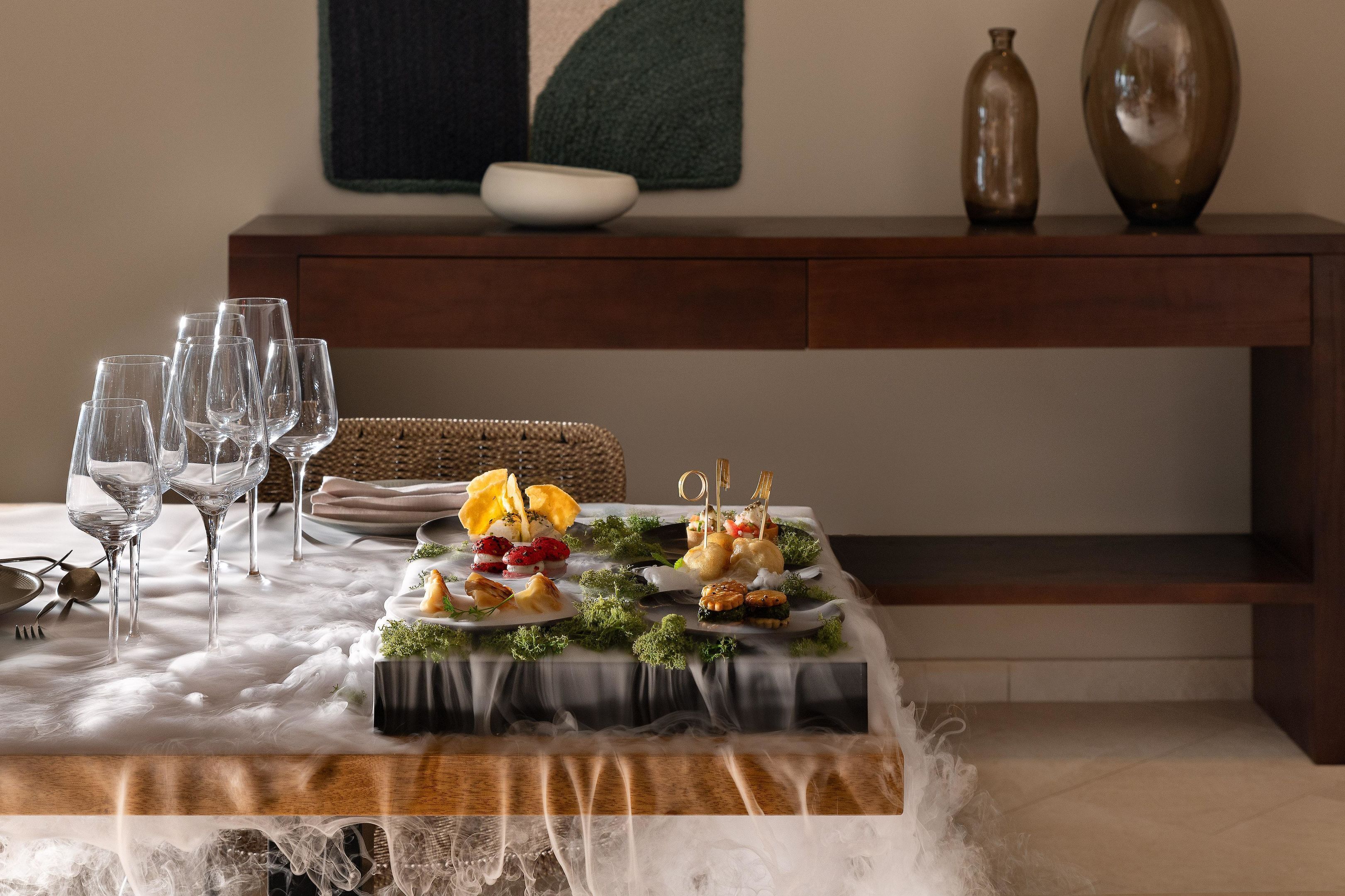 Una mesa de comedor dispuesta con vasos transparentes, platos y una variedad de aperitivos.