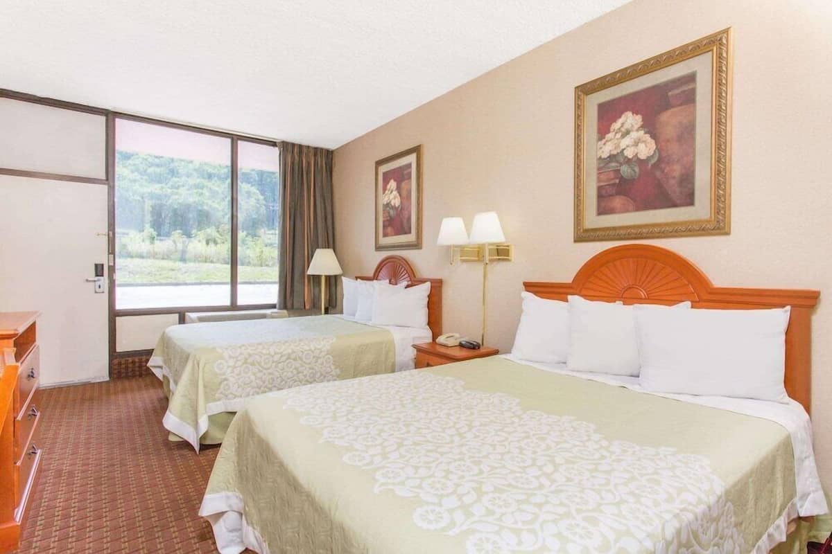 A hotel room with two beds, a large window with a view of greenery, a desk, and a lamp.