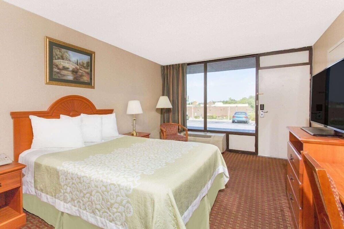 A hotel room with a bed, a television, a chair, a lamp, a painting on the wall, and a window with a view of a parking lot.