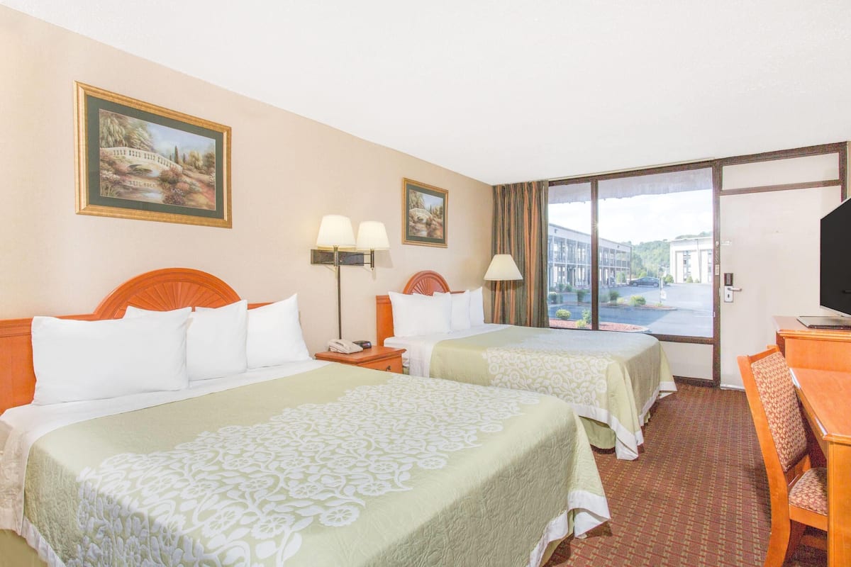 A hotel room with two beds, a desk, a TV, and a large window offering a view of buildings and a parking lot.