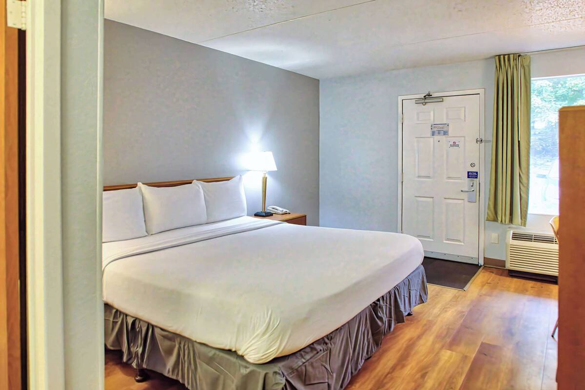 A neatly made bed with a white comforter and a wooden headboard. There is a bedside table with a lamp and a phone. A door with a sign is visible in the background.