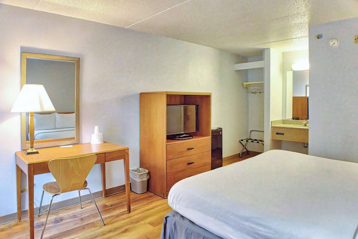 A hotel room with a wooden desk, a chair, a television, a bed, a mirror, and a bathroom with a sink and mirror.