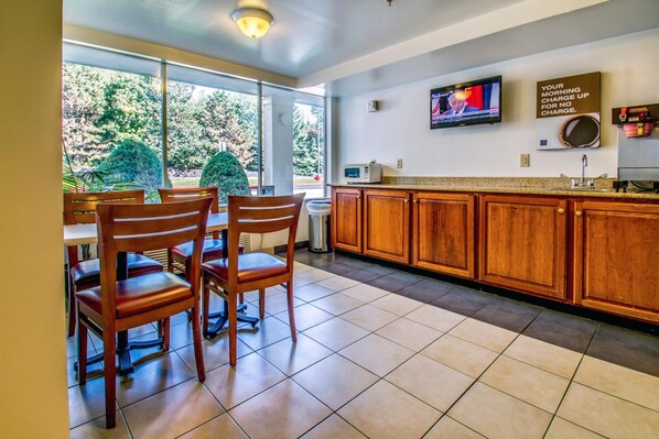 A modern kitchen with wooden cabinets, a dining area with chairs, a television, and a large window with a view of trees.