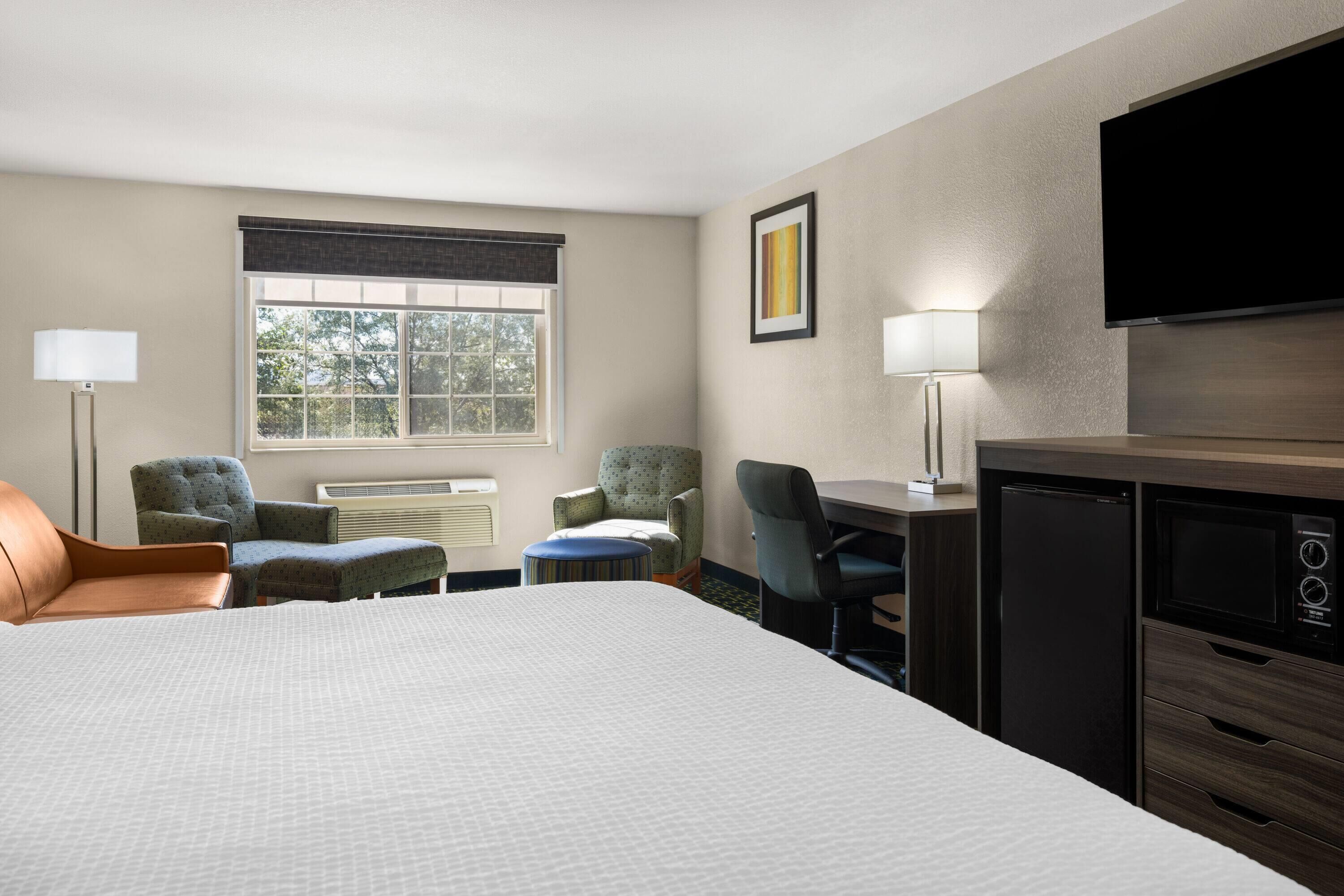 A hotel room with a bed, two armchairs, a desk, a television, and a kitchenette.