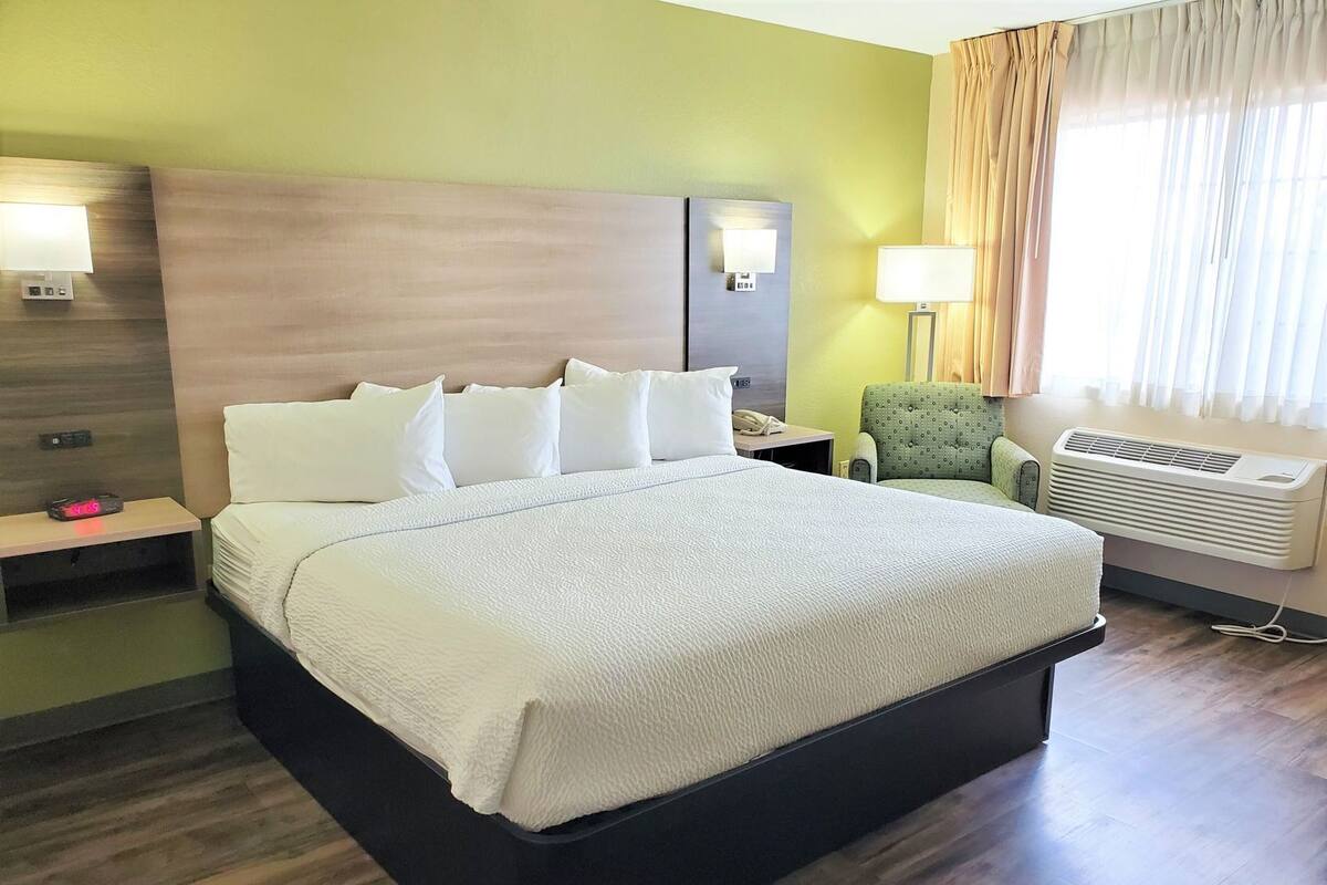 A hotel room with a large bed, bedside tables, a chair, a lamp, and an air conditioning unit.