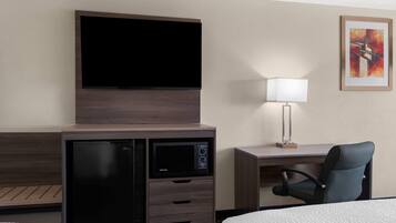 A hotel room with a flat-screen TV mounted on a wooden cabinet, a desk with a lamp, and a chair.