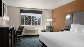 A hotel room with a bed, desk, chair, air conditioning unit, and a window with a view of trees.