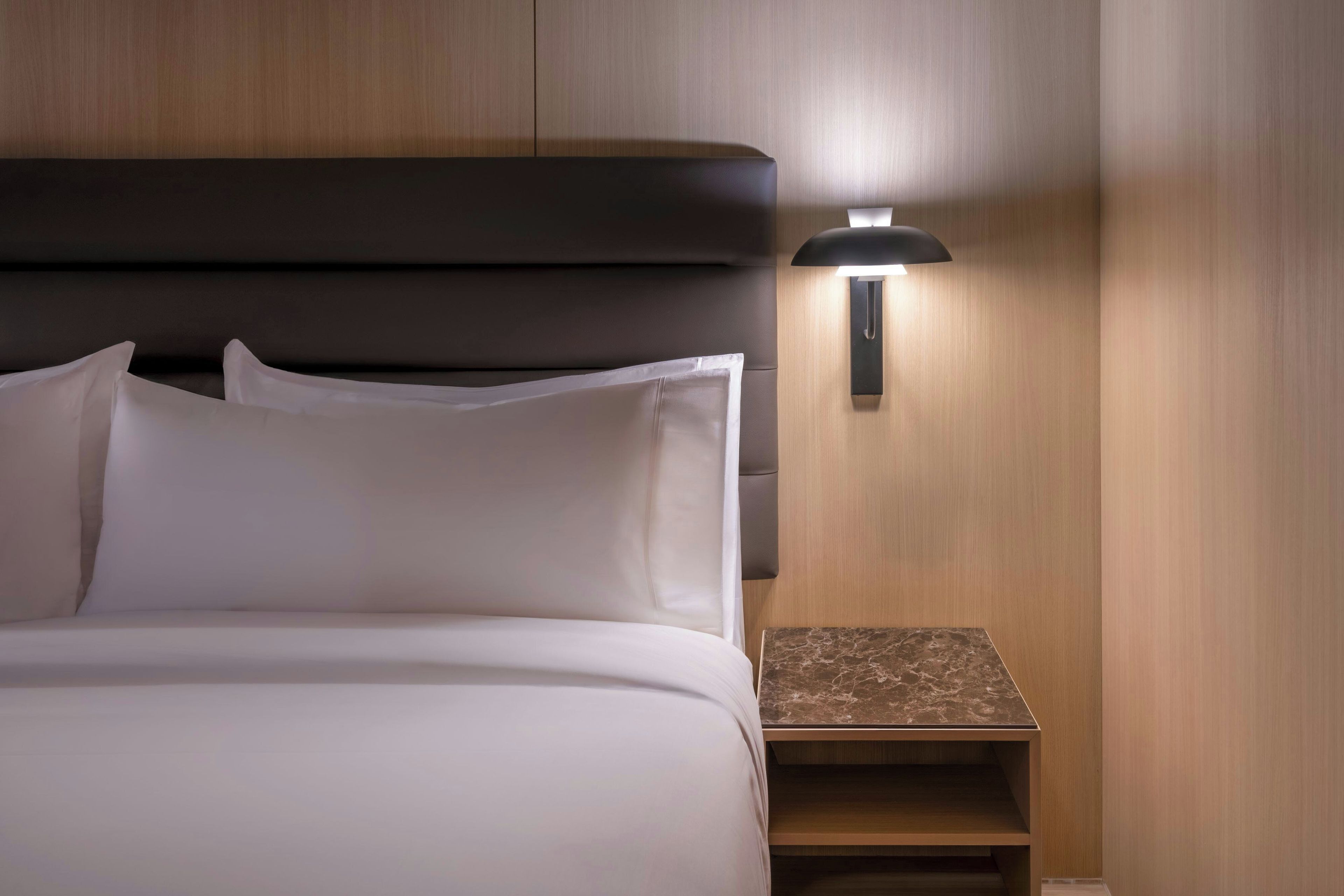 A neatly made bed with white linens and a headboard, a bedside table with a marble top, and a wall-mounted lamp.