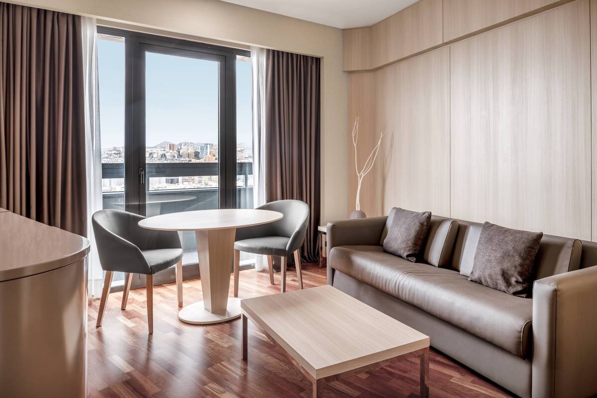 Una sala de estar moderna con un sofá, dos sillones, una mesa redonda y vistas a un paisaje urbano a través de amplios ventanales.