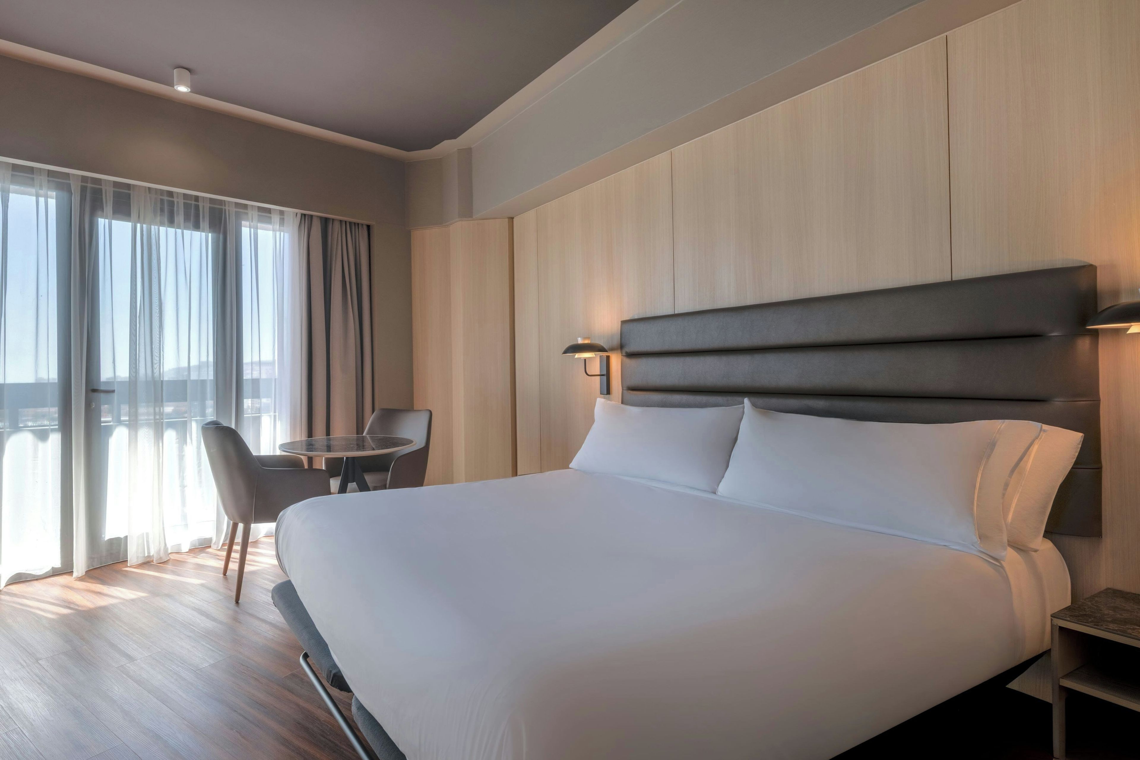 Une chambre d’hôtel avec un grand lit, un bureau, une chaise et une vue sur la ville par les fenêtres.