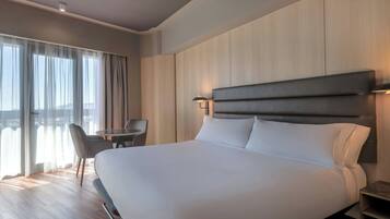 Une chambre d’hôtel avec un grand lit, un bureau, une chaise et une vue sur la ville par les fenêtres.