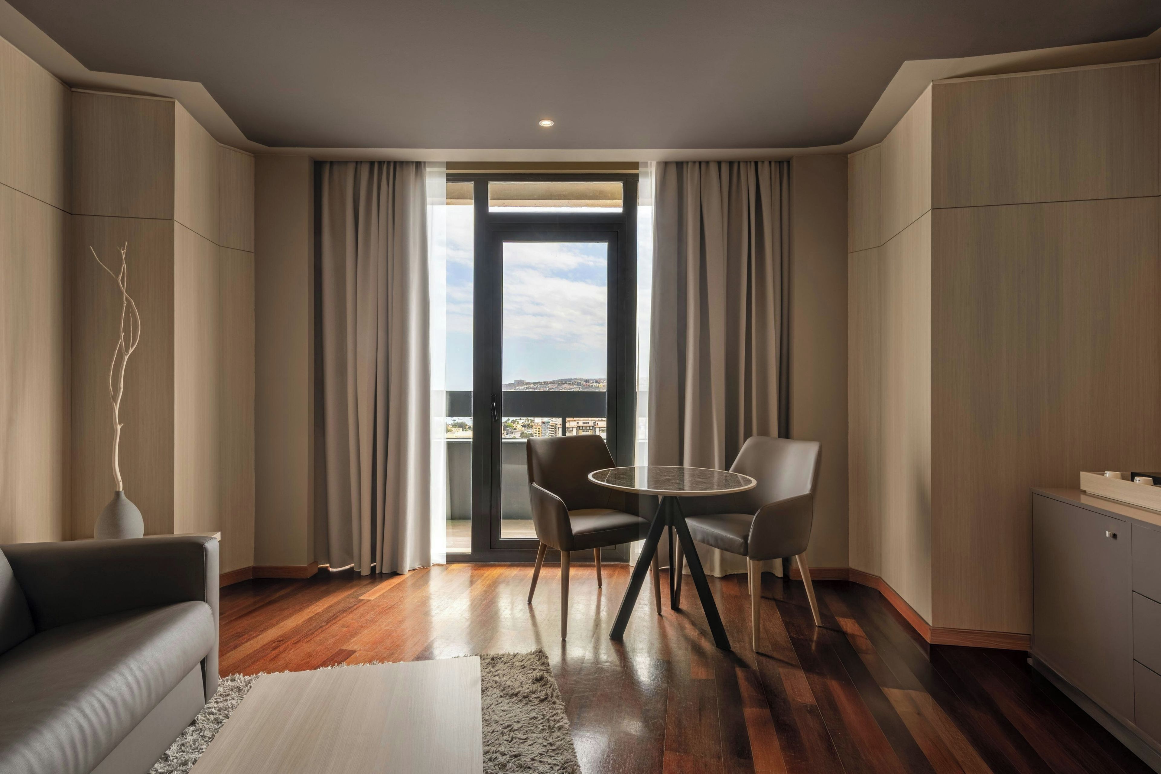 Una sala de estar moderna con una mesa de vidrio, dos sillas, un sofá y vistas a través de un ventanal.