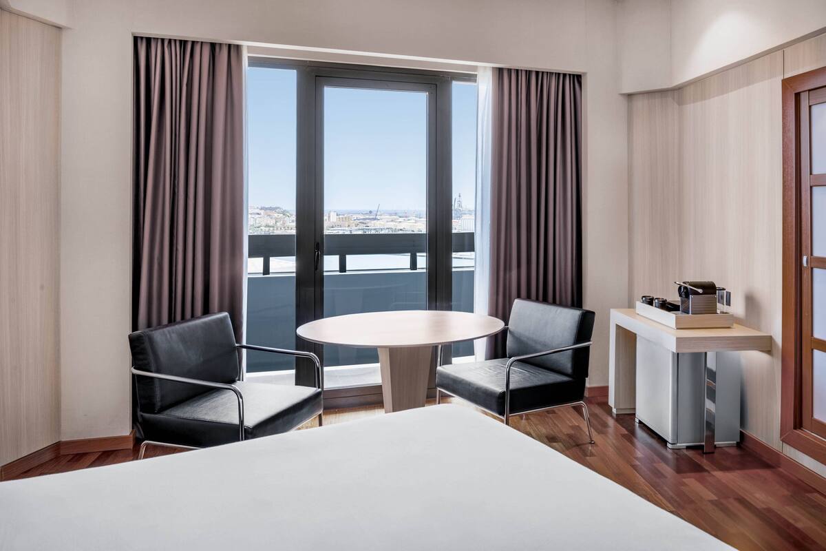 Habitación de hotel con una cama grande, dos sillones, una mesita redonda y un escritorio con silla. Cuenta con un ventanal con cortinas y vistas a un paisaje urbano y un puente.