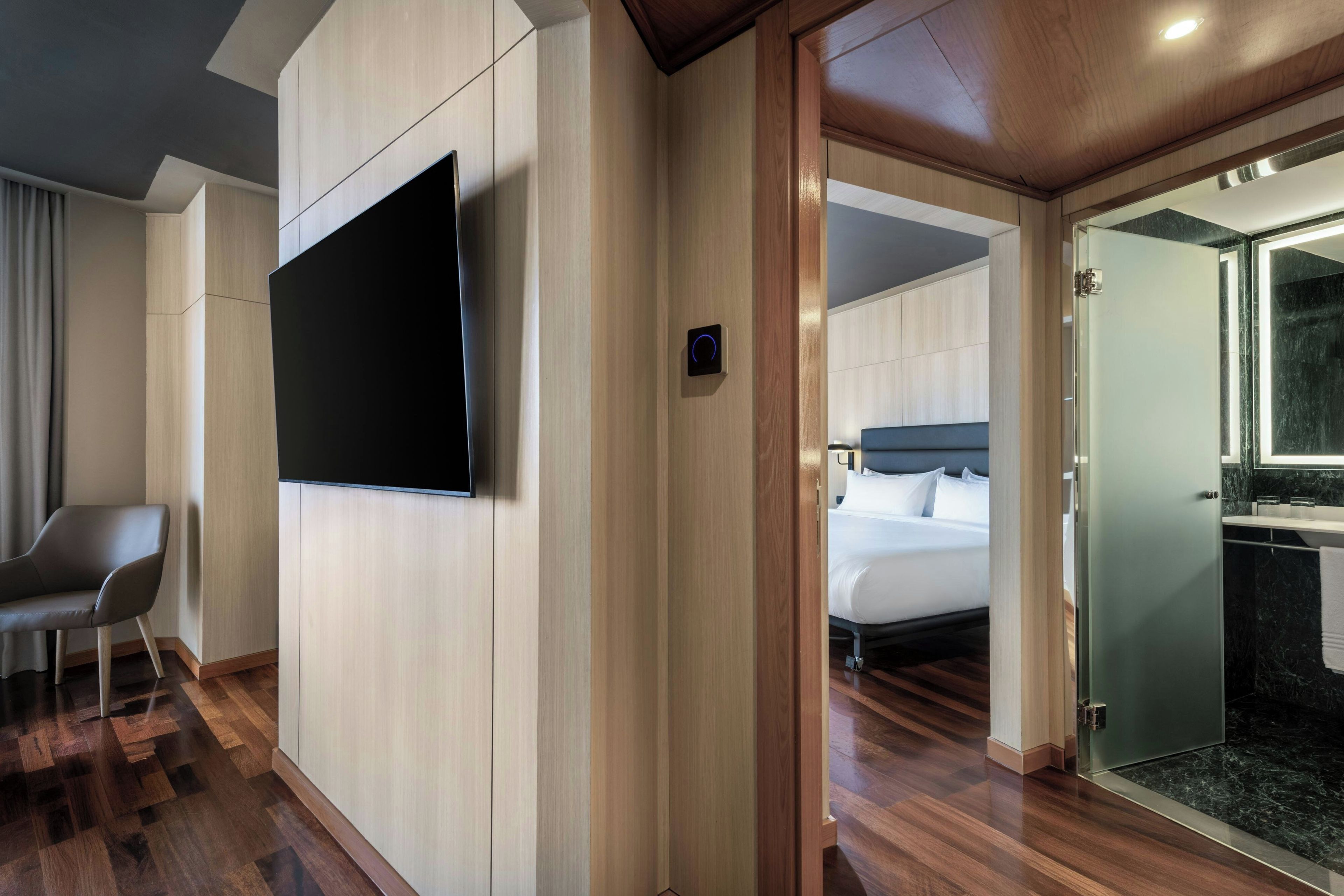 Habitación de hotel con televisor de pantalla plana montado en la pared, cama con cabecera y baño con cabina de ducha de vidrio.