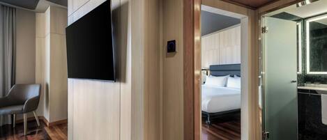 Habitación de hotel con televisor de pantalla plana montado en la pared, cama con cabecera y baño con cabina de ducha de vidrio.