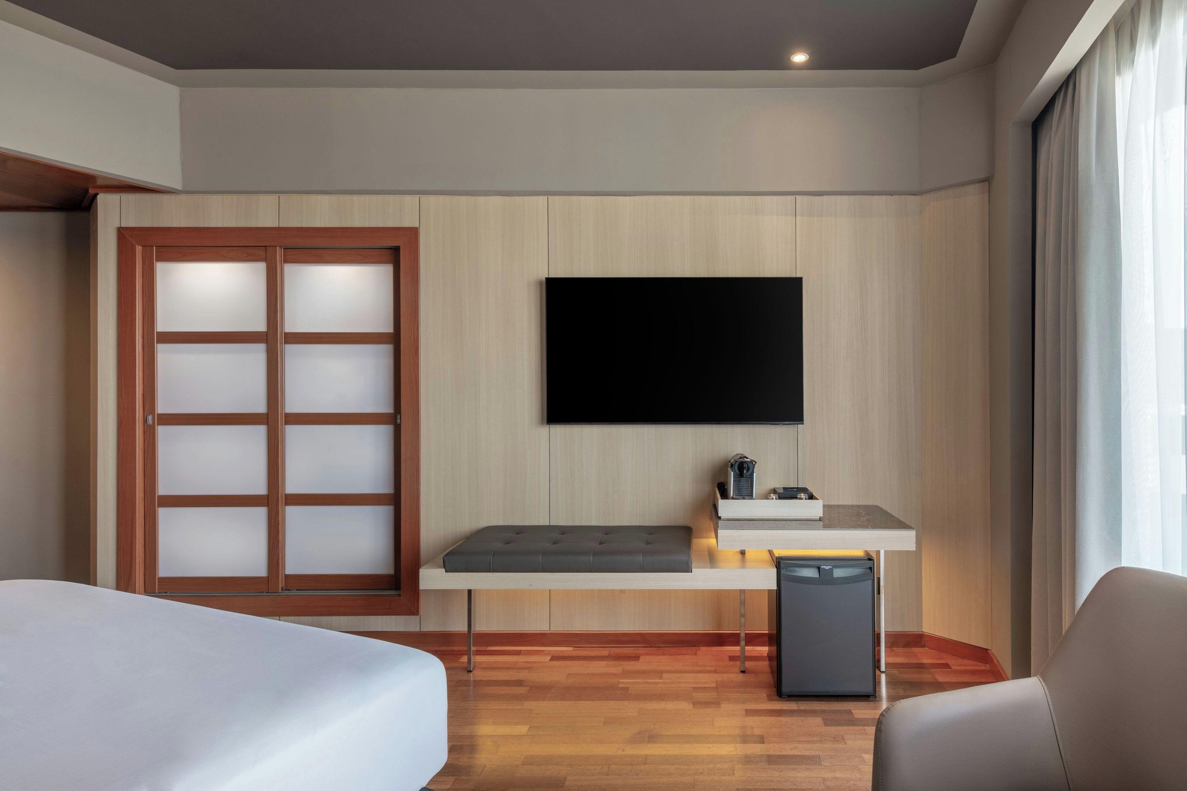 Una camera d'albergo con un letto, una scrivania con TV a schermo piatto, un'area salotto e un armadio con pannelli trasparenti.