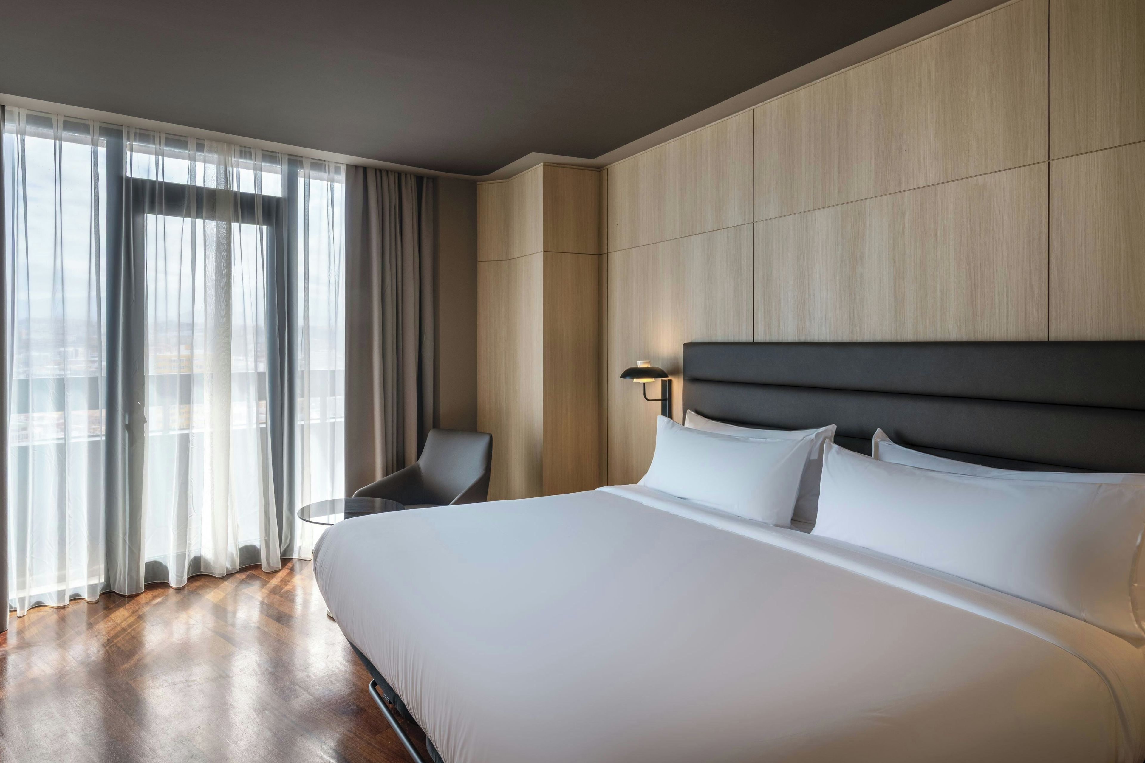 A hotel room with a large bed, a chair, a small table, and a window with curtains.