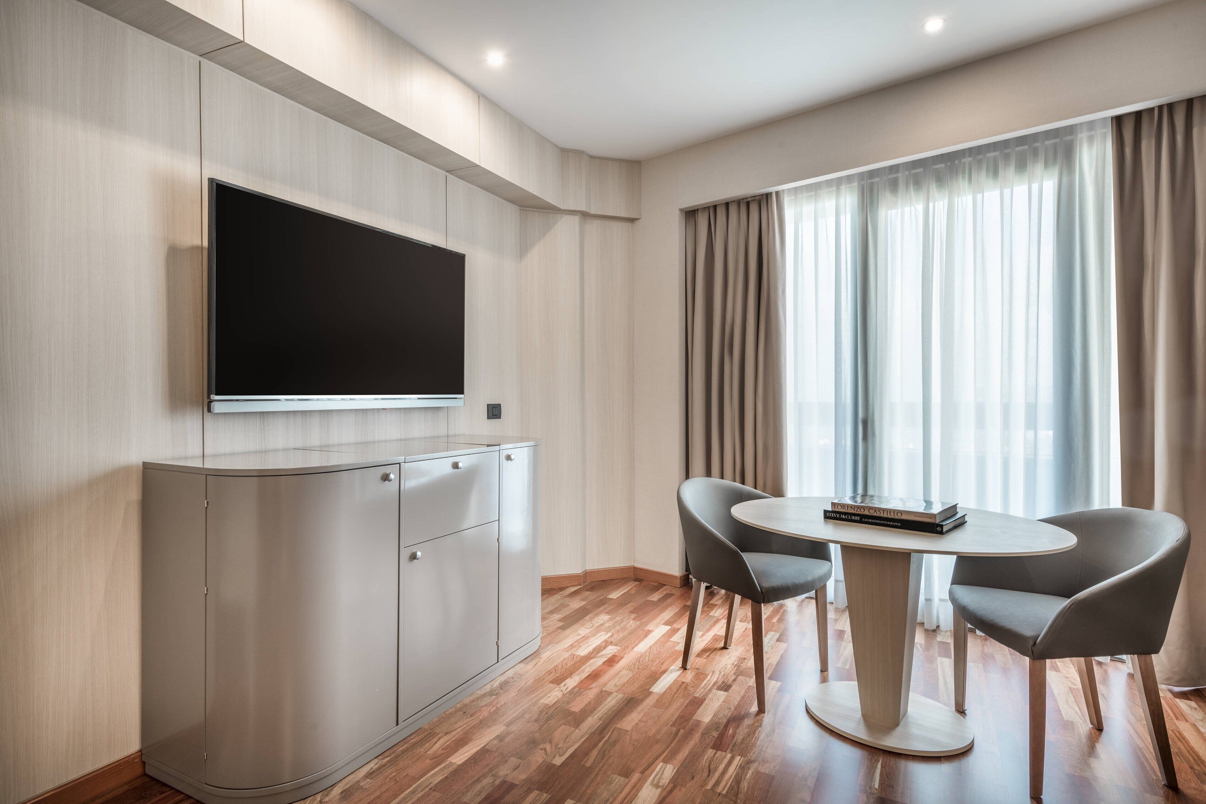 Una habitación de hotel moderna con un televisor de pantalla plana montado en un mueble, una mesa redonda con dos sillas y un ventanal con cortinas.