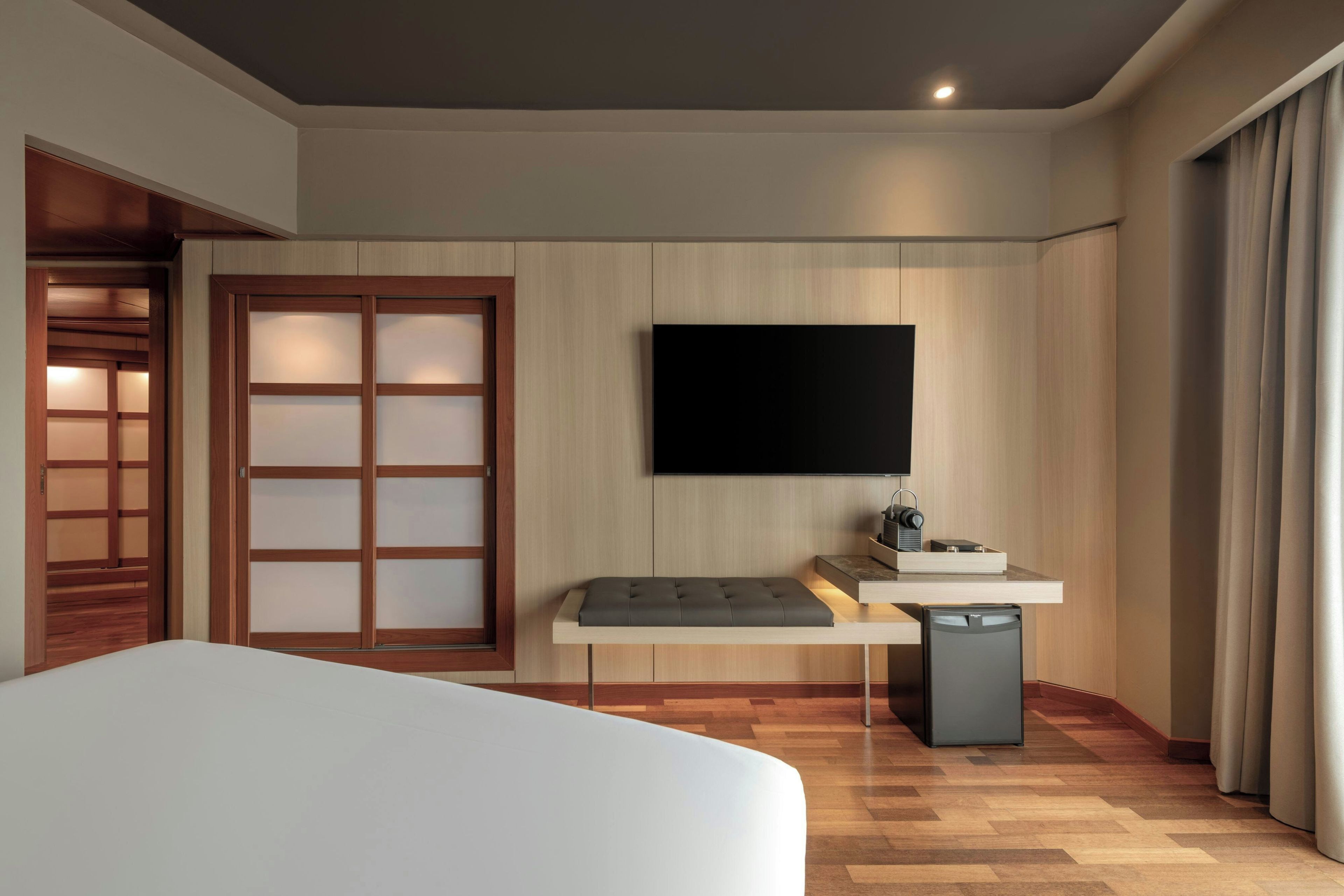 A modern hotel room with a bed, a flat-screen TV mounted on the wall, a small bedside table with a lamp, and a wooden wardrobe with a sliding door.