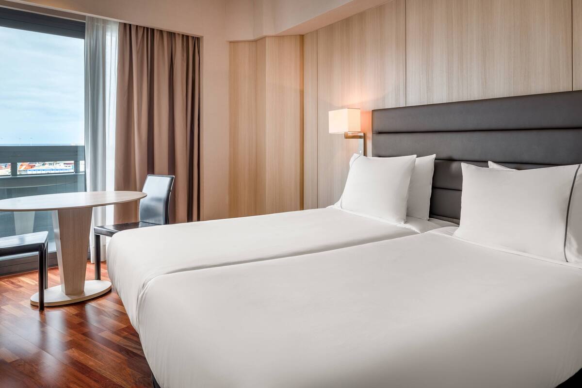 Habitación de hotel con dos camas, una mesita, una silla y vistas a la ciudad.