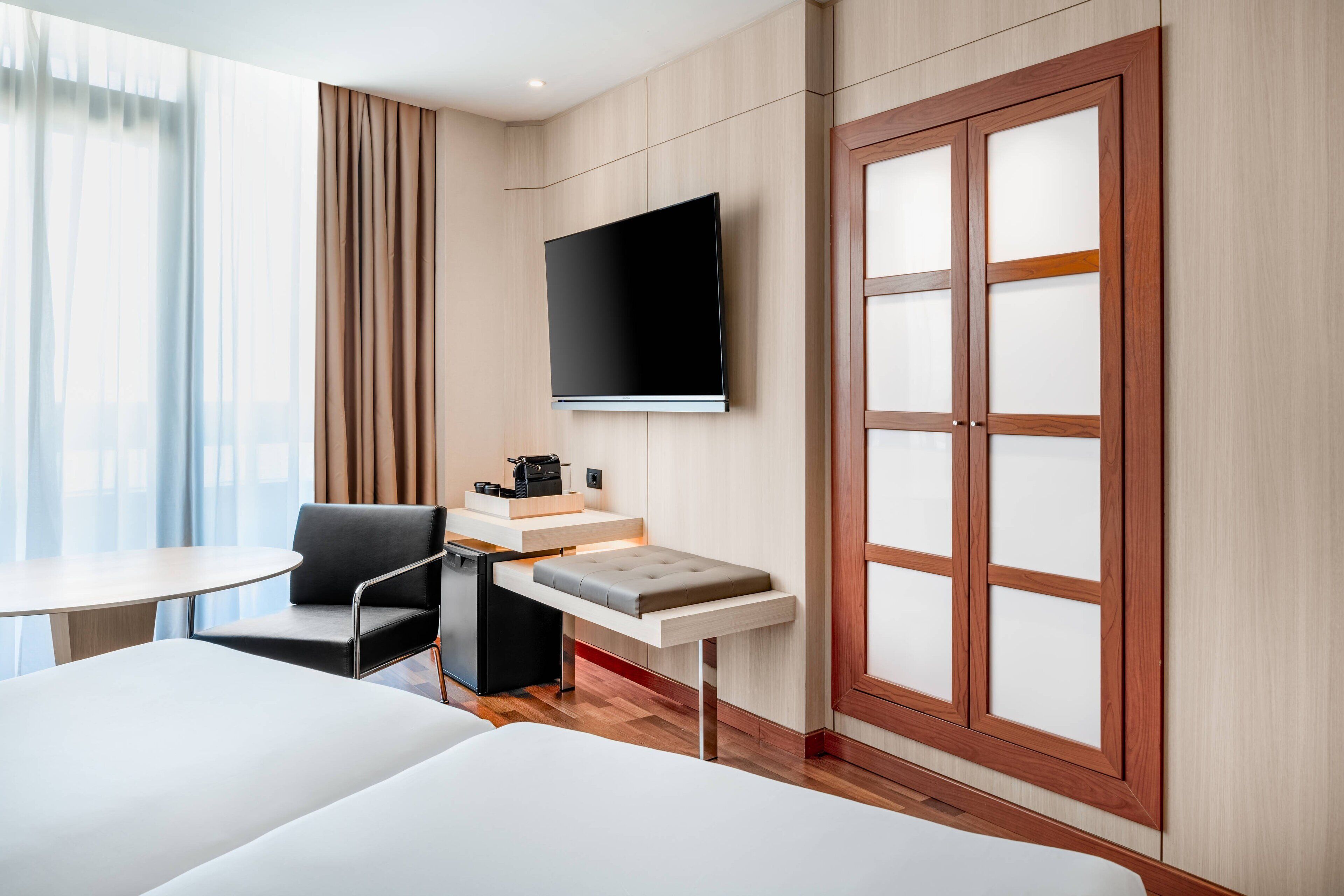 Une chambre d’hôtel comprenant un lit, un bureau, une chaise, un téléviseur fixé au mur et une grande fenêtre avec des rideaux.