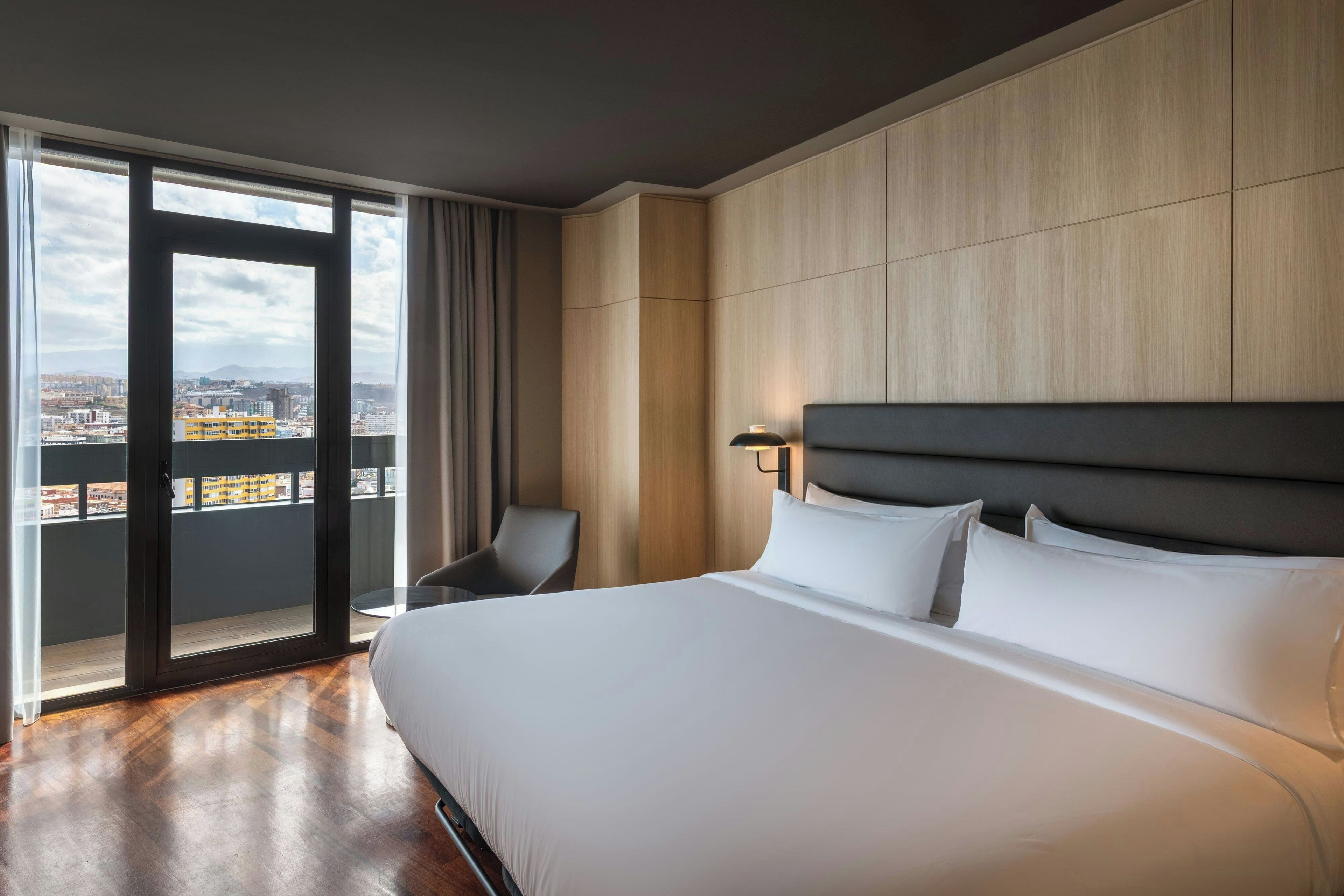Camera d'albergo con un letto grande, una poltrona, un comodino e vista sulla città tramite un balcone.