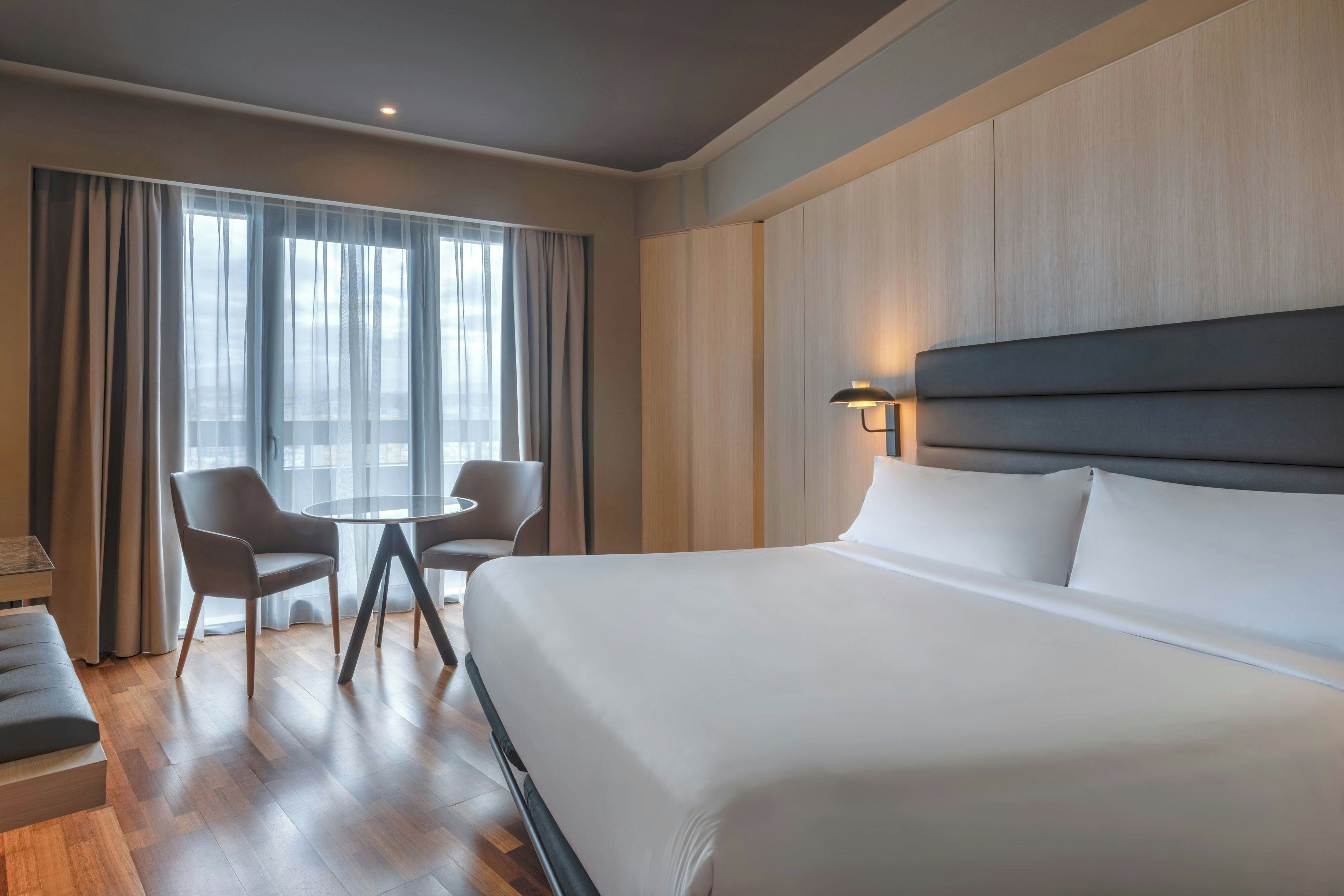 A hotel room with a large bed, a round table with two chairs, and a view through sheer curtains.