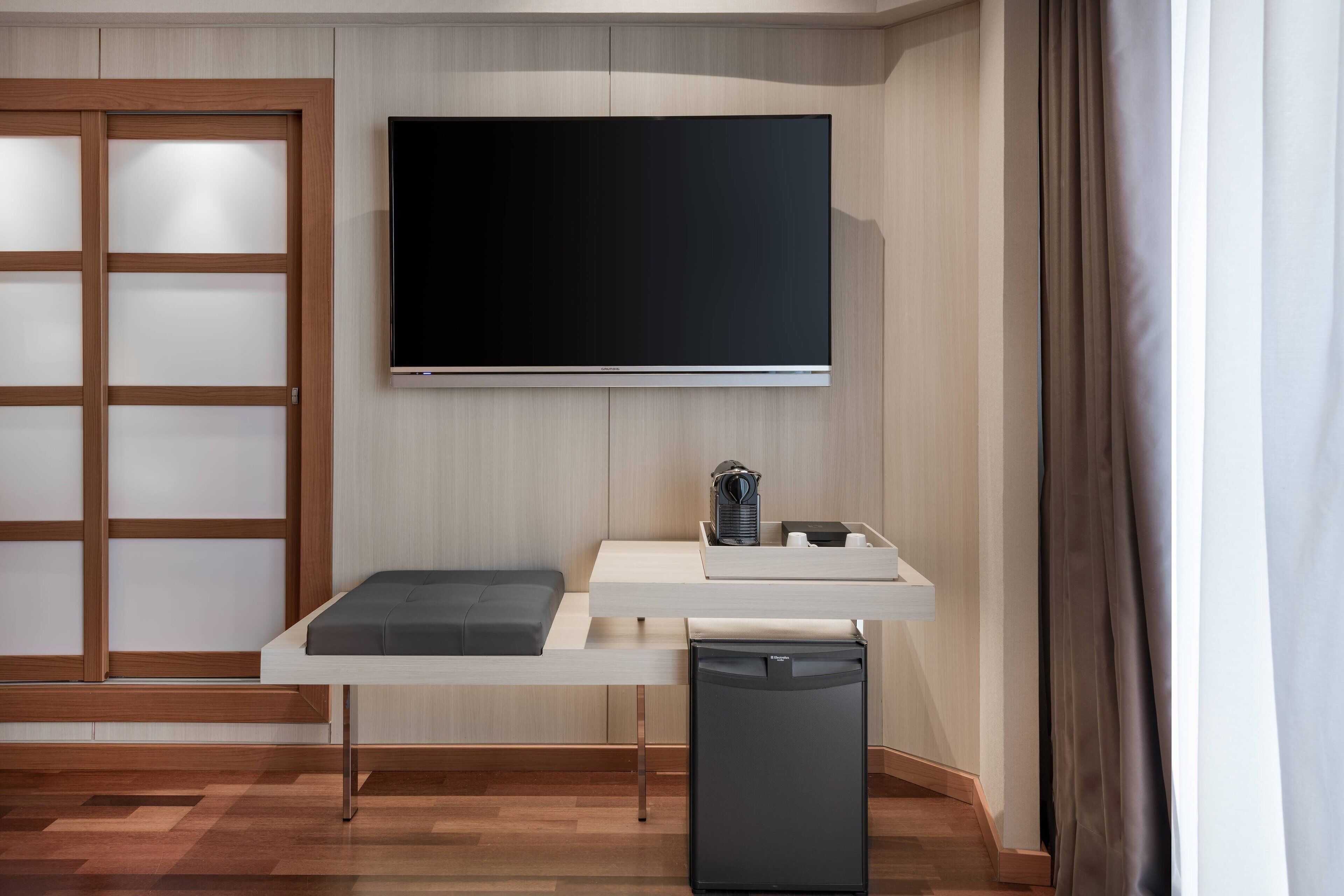 A modern room with a flat-screen TV mounted on the wall, a small table with a bench, and a compact refrigerator.