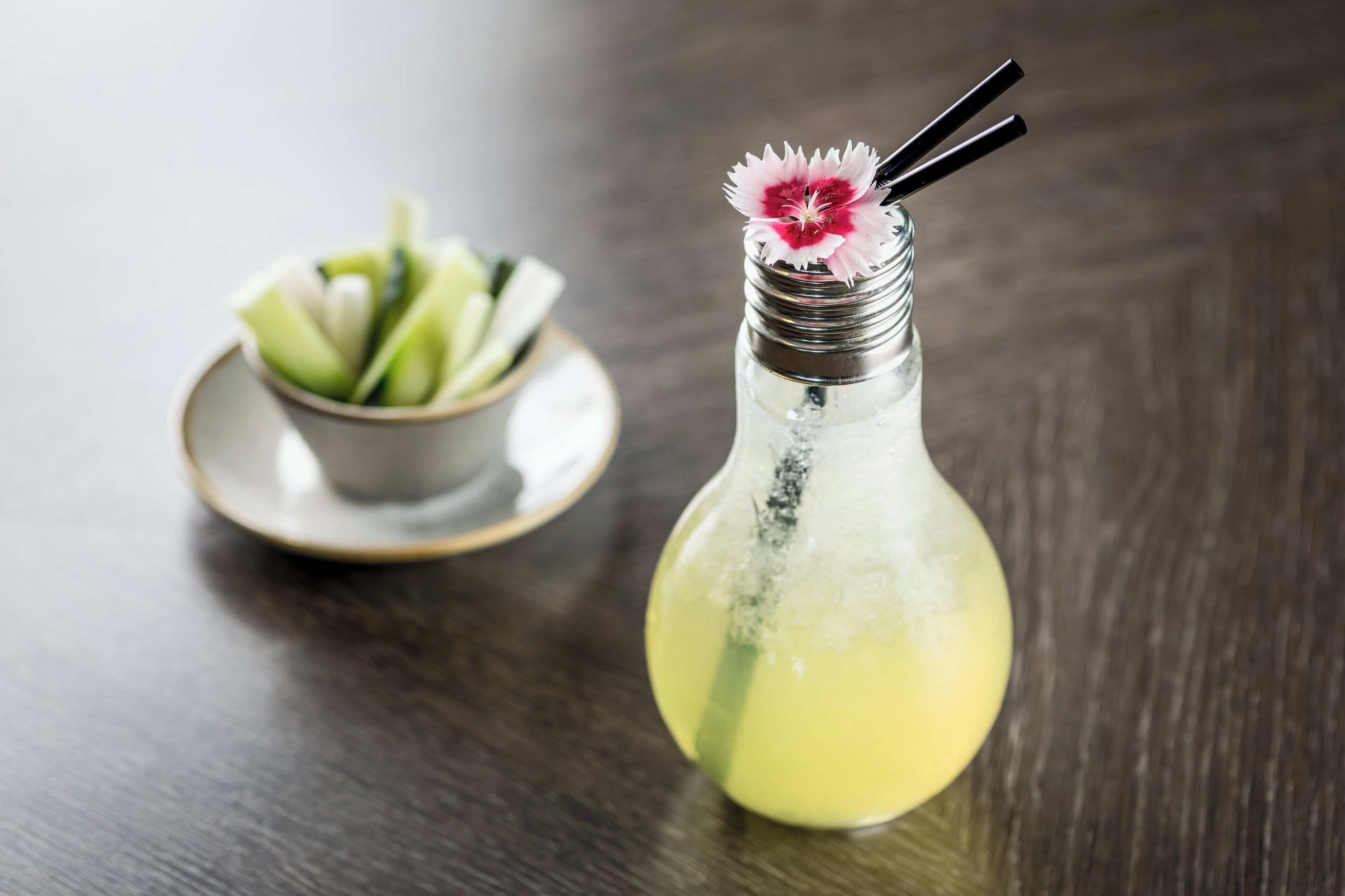 Un cóctel en una botella de vidrio, adornado con una flor y una pajita, acompañado de un cuenco pequeño con fruta cortada sobre un platito.