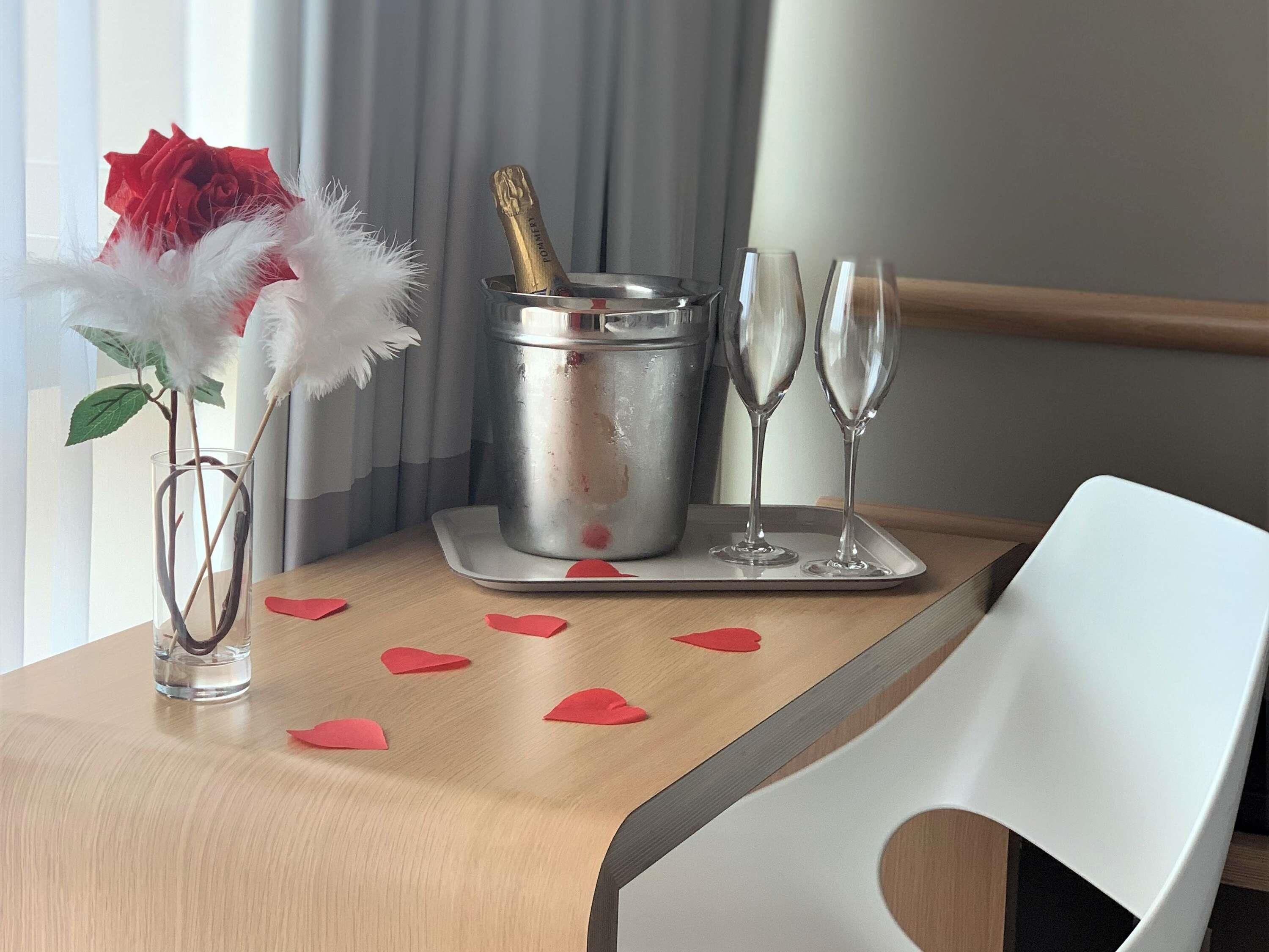 A table with a vase of a red rose, a champagne bottle, two champagne flutes, and red paper hearts on a wooden table.