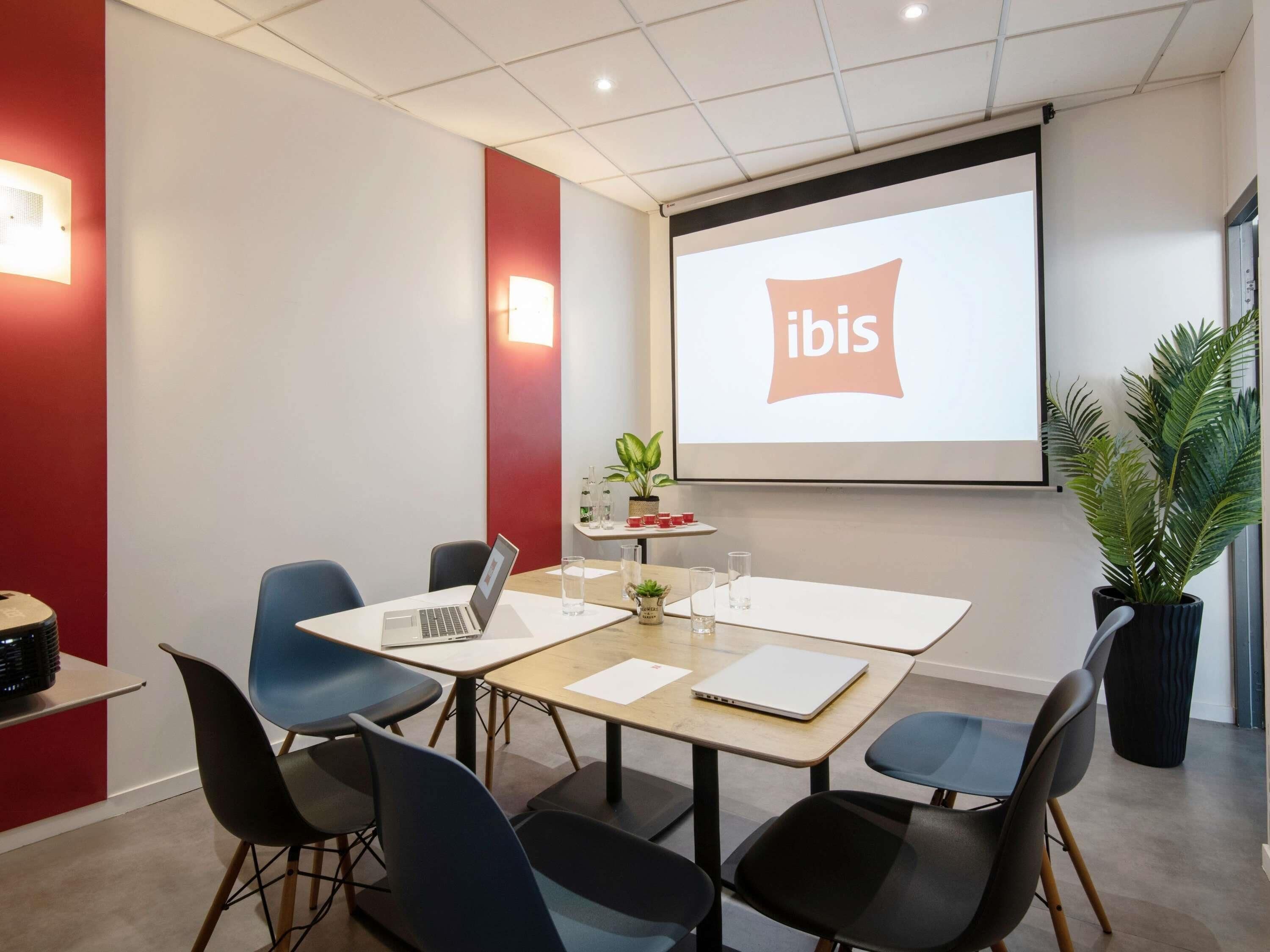 Sala de conferências com tela de projetor exibindo 'ibis', uma mesa com laptops, cadeiras, uma pequena planta e um bebedouro.
