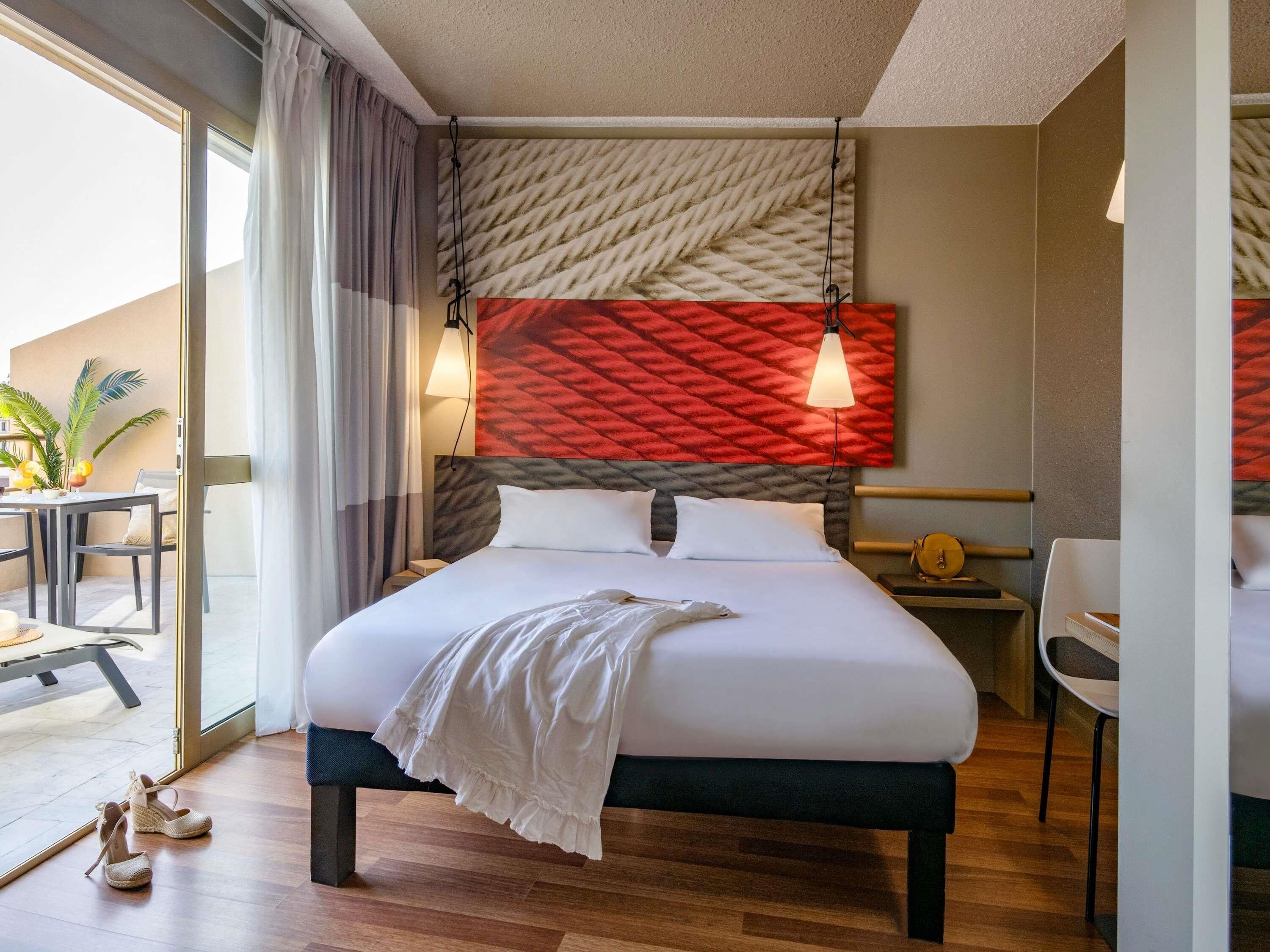 Quarto de hotel com cama, escrivaninha e cadeira. Tem uma janela com cortinas, uma planta e vista para o exterior.