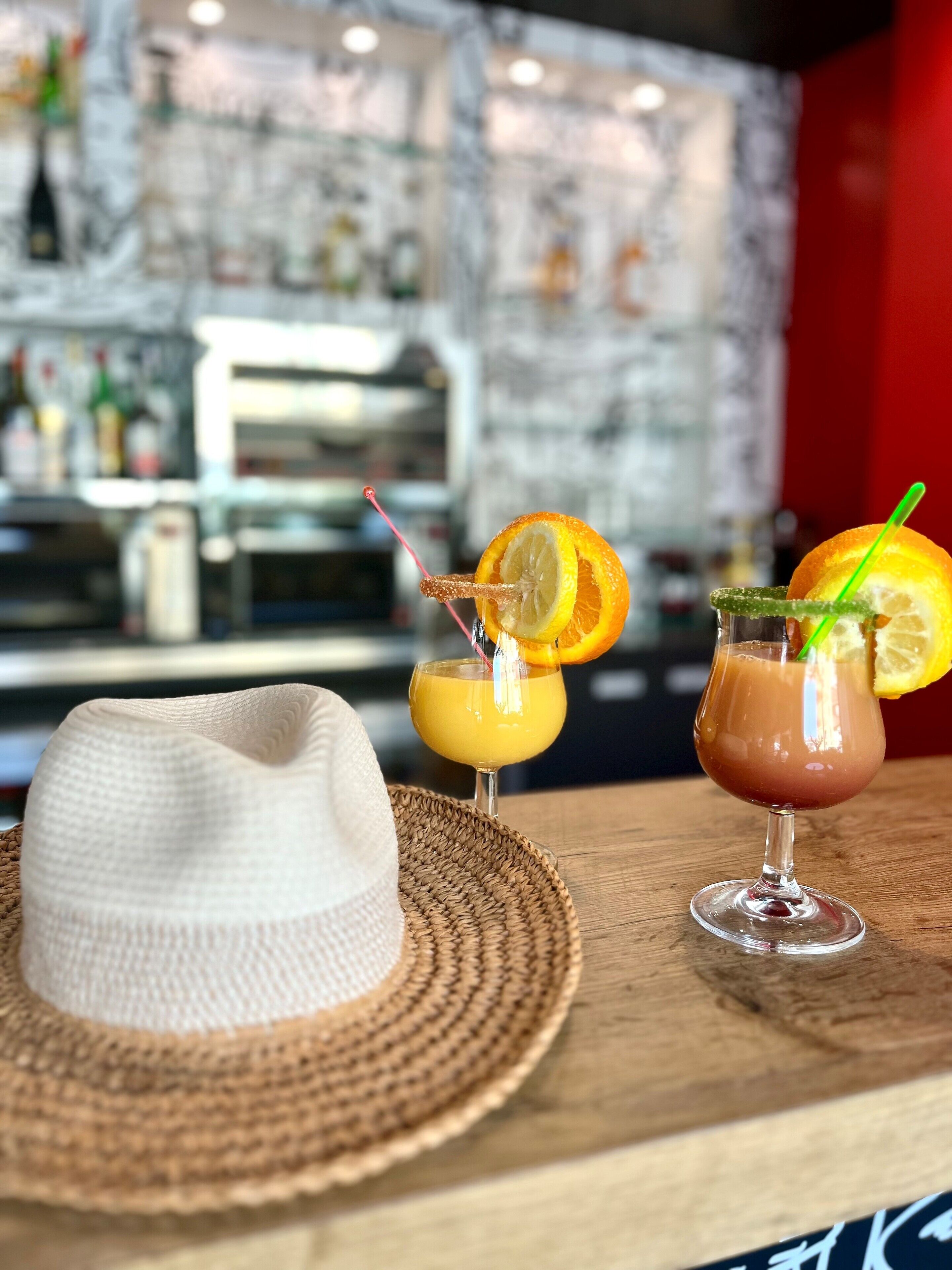 A bar area with a hat on a woven mat, two cocktails with garnishes, and a blurred background with bottles.
