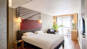 A hotel room with a large bed, bedside lamps, a desk with a computer, a TV, and a window with curtains.