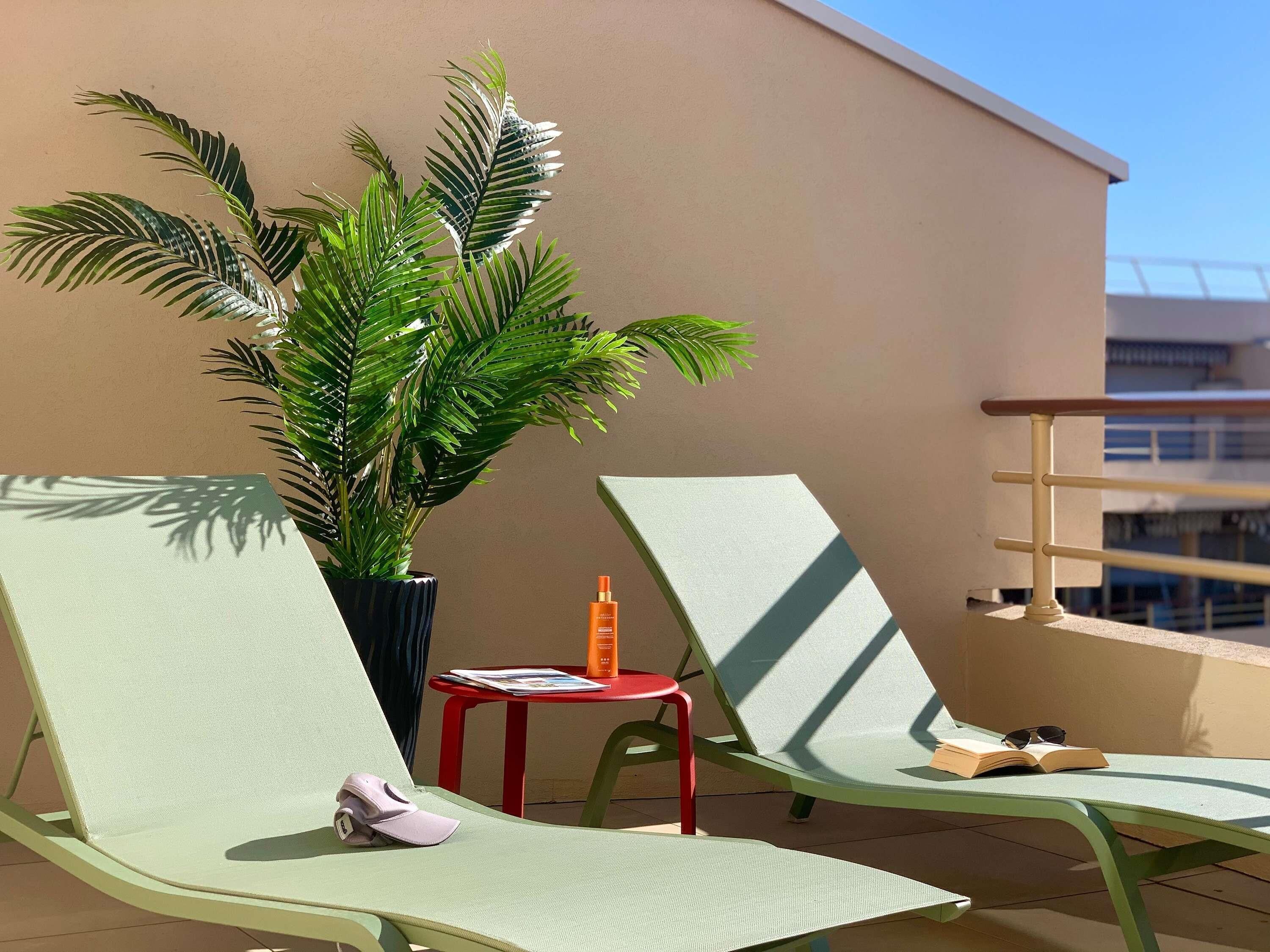 A balcony with two green lounge chairs, a small red table with a bottle and a book, and a potted plant.