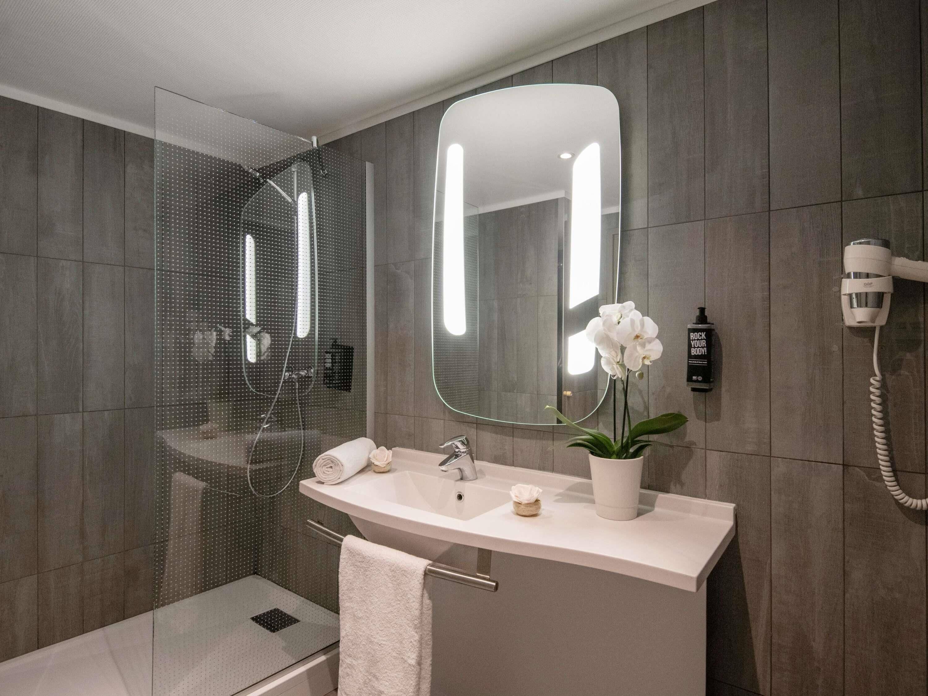 Ein modernes Badezimmer mit einer ein Glasfach rahmenden Dusche, einem großen Spiegel, einem Waschbecken mit Topforganide und einem an der Wand befestigten Föhn.