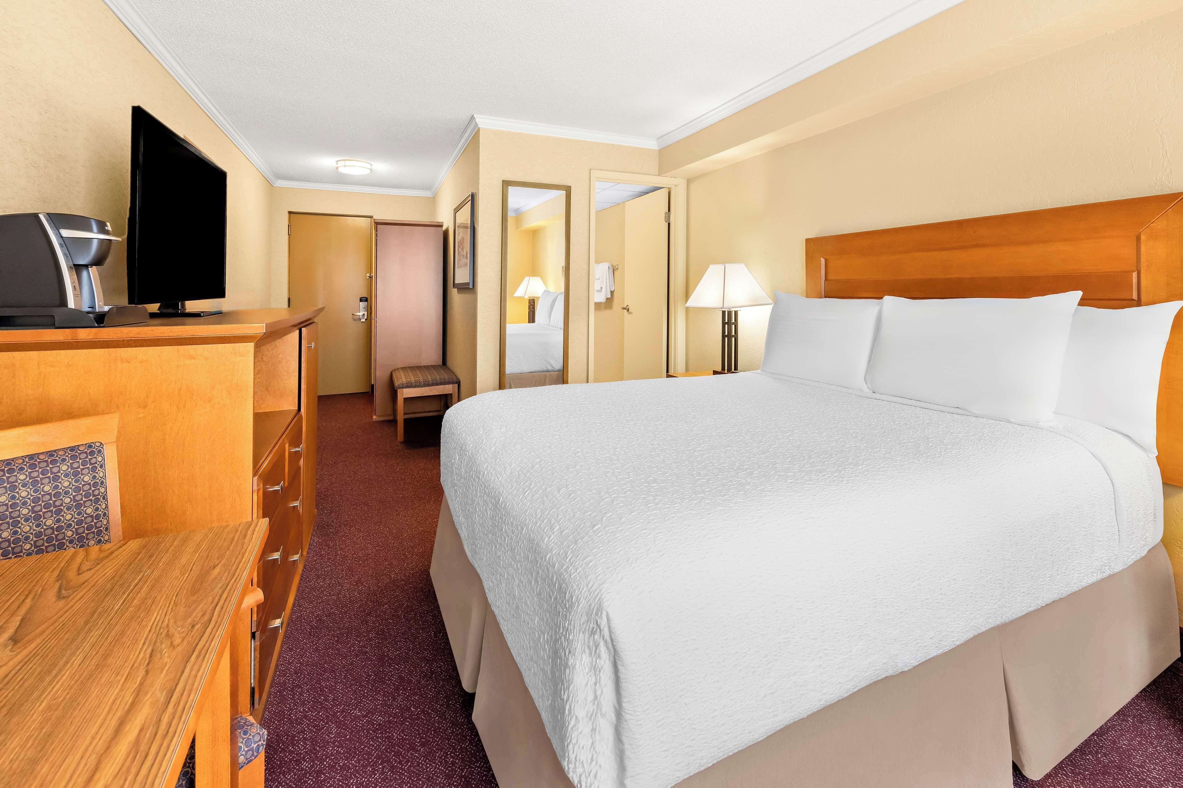 Habitación de hotel con una cama grande, un escritorio con computadora y cafetera, una silla y una puerta que da a otra habitación.