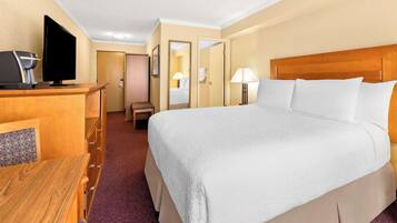 Habitación de hotel con una cama grande, un escritorio con computadora y cafetera, una silla y una puerta que da a otra habitación.