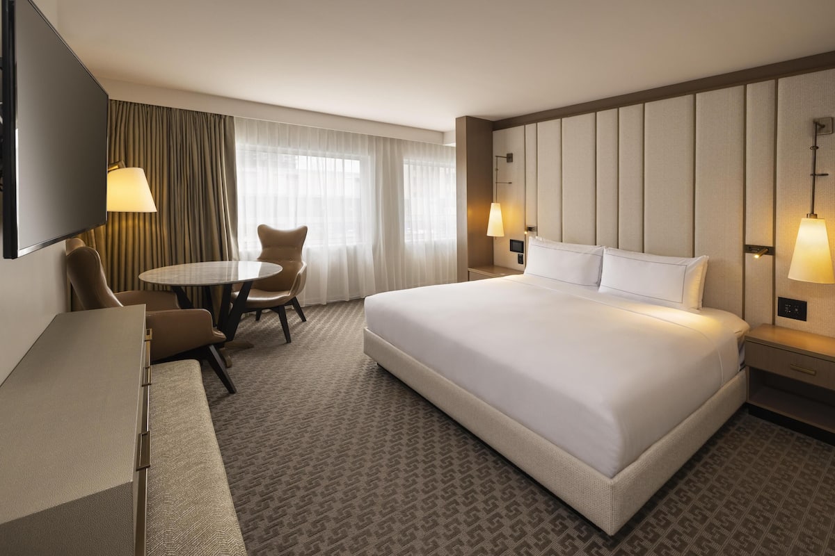 A hotel room with a large bed, a desk, a chair, a small table, and two lamps.