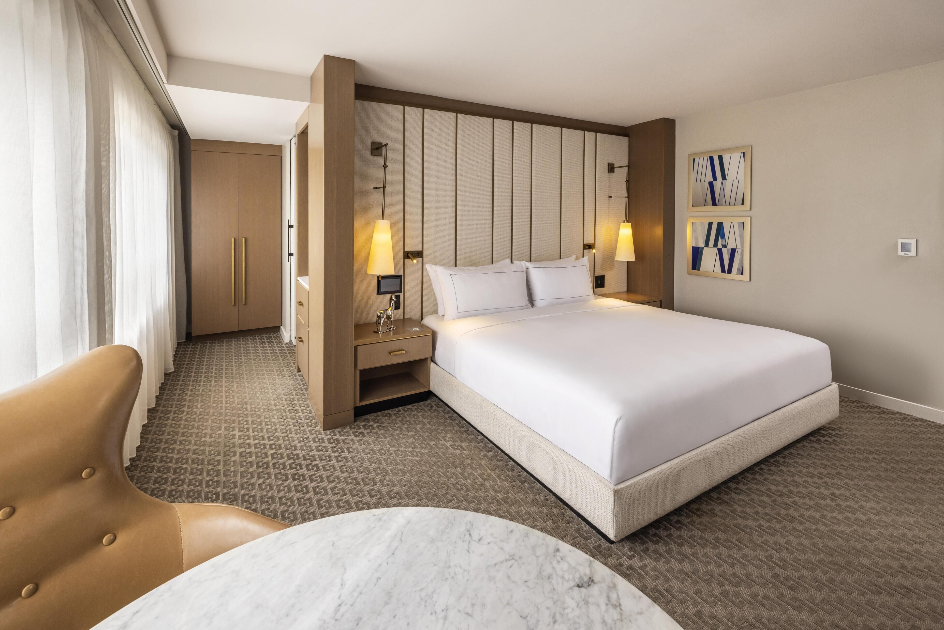 A hotel room with a large bed, a desk with a lamp, a chair, and a marble table.