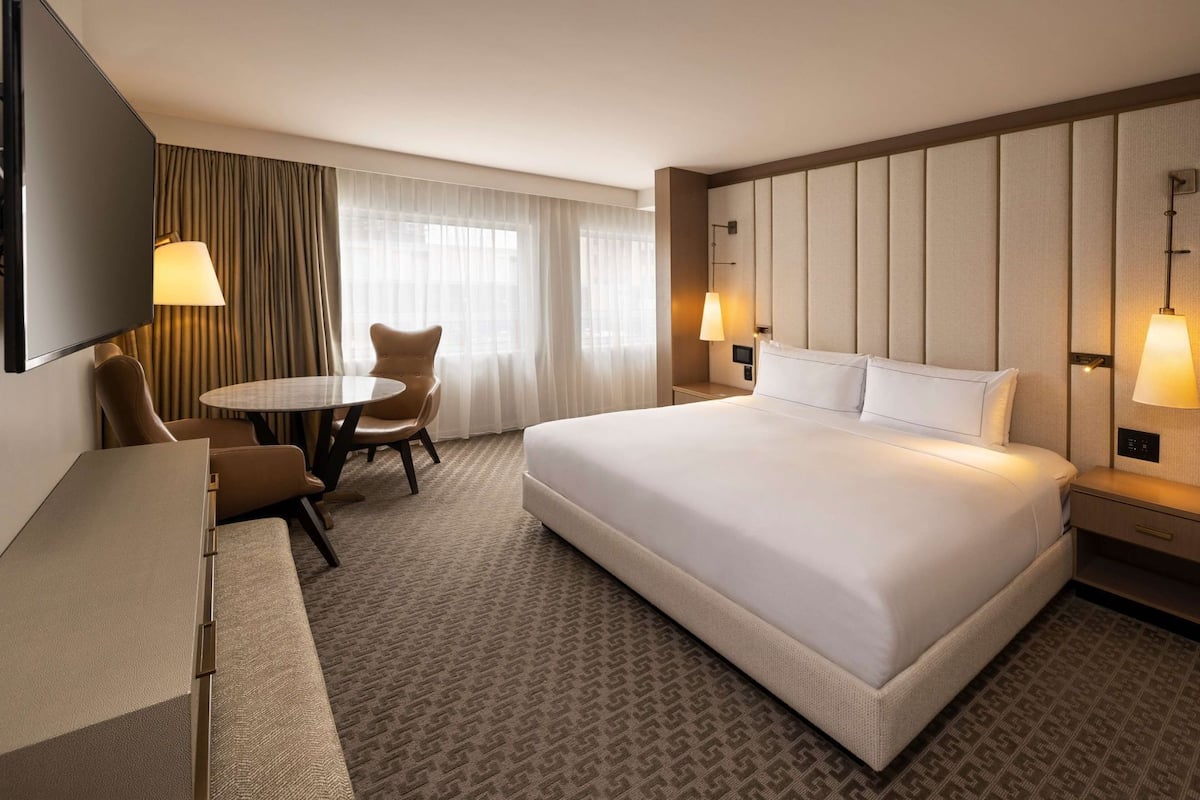 A hotel room with a large bed, a desk, a chair, a small table, and a TV.