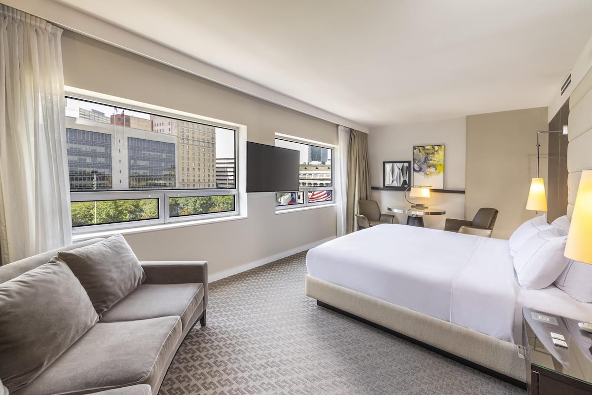 A hotel room with a large bed, a sofa, a TV, a desk with a lamp, and large windows with a city view.